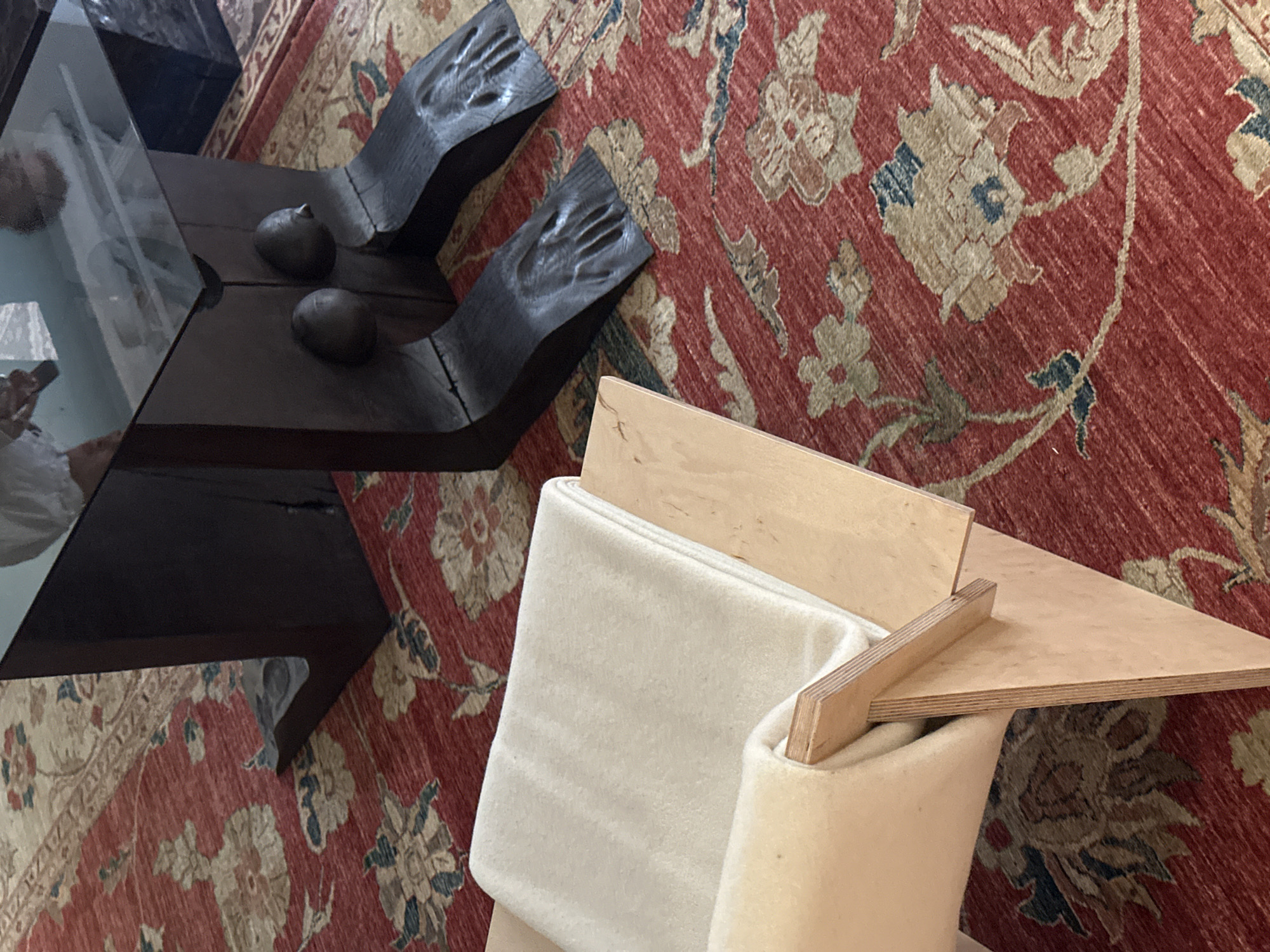A high-angle shot displays an oriental rug on which a dark brown coffee table with a glass top and hand-shaped legs sits next to a light cream chair with plywood accents. The environment appears to be indoors. The location in London, United Kingdom, cannot be determined from this image.