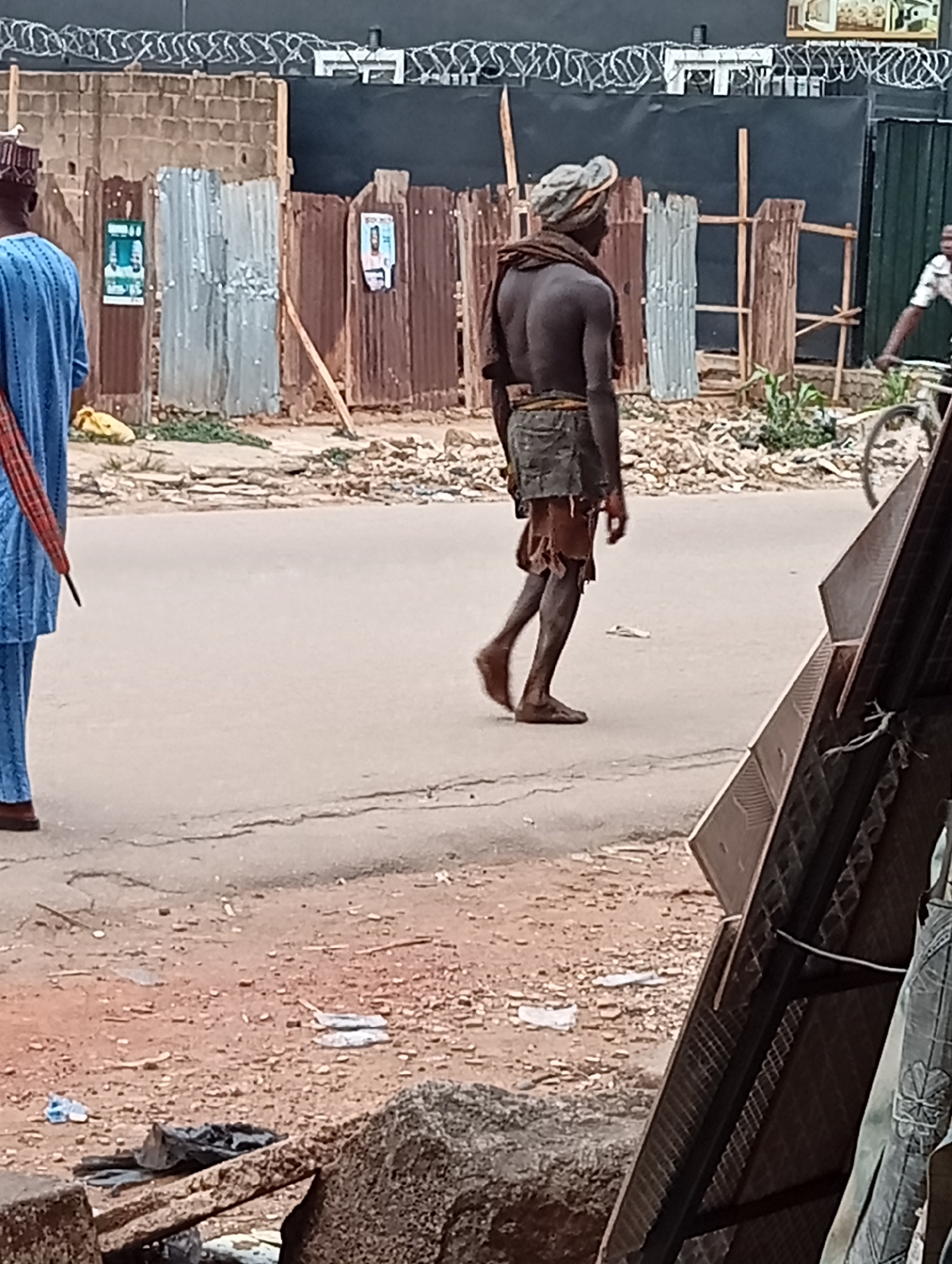 The image shows a street scene in Maiburiji, Nigeria. A man with bare torso and wearing torn shorts walks barefoot down a paved road. He has a head covering and a dark cloth draped around his neck. To the left is a man wearing a blue garment, facing away from the camera. In the background, there are makeshift fences made of corrugated metal sheets and wooden supports. A person on a bicycle is partially visible on the right side of the frame. In the foreground, there is a large metal object and debris on the side of the road. Razor wire tops a brick wall.