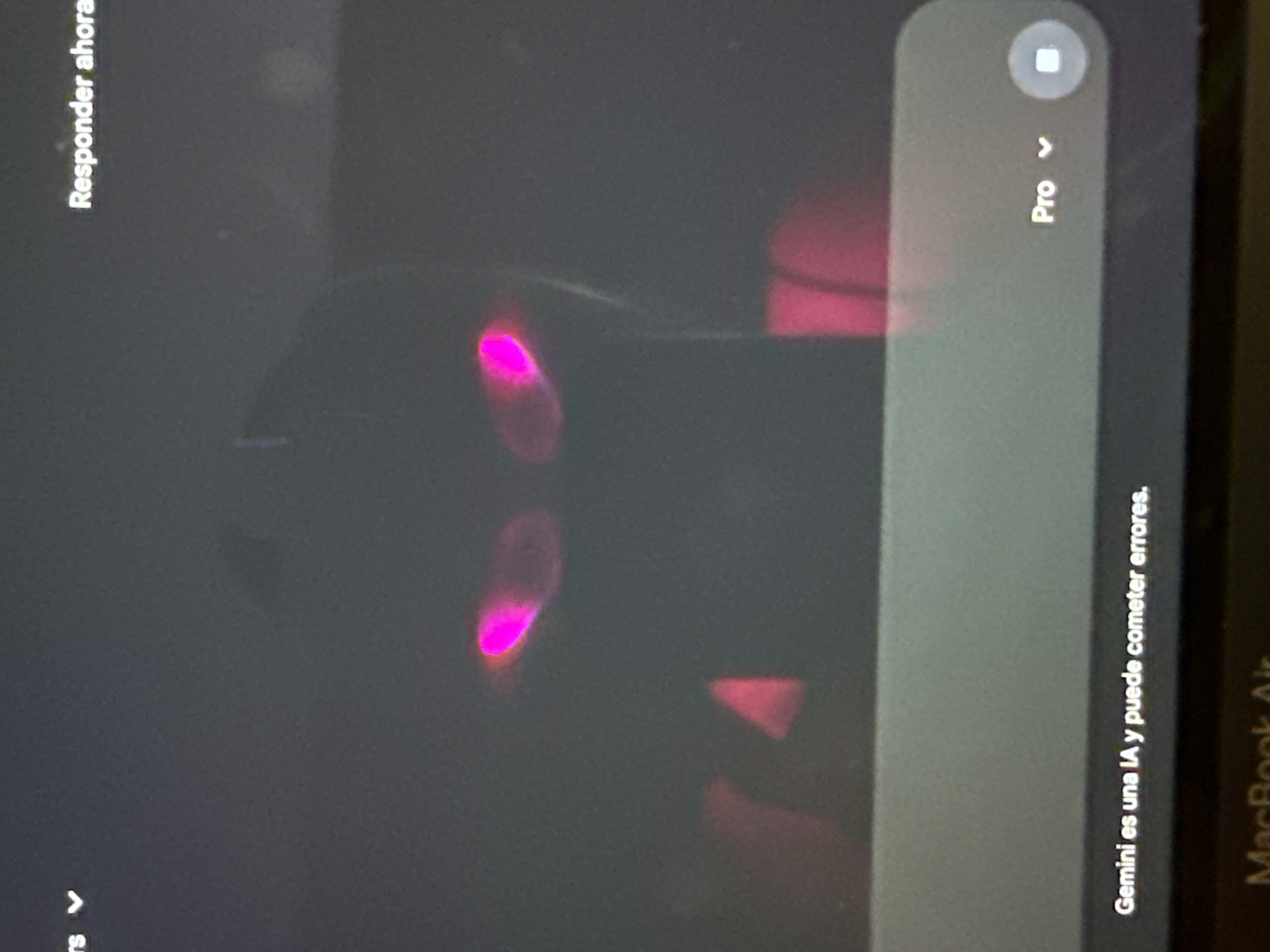 The image, taken in San Bernardino Tlaxcalancingo, Mexico, captures a dimly lit screen, likely a laptop given the faint "MacBook Air" text visible on the right edge. The screen displays a dark digital interface, oriented vertically, with text rotated.
On the left side, rotated 90 degrees counter-clockwise, the text "Responder ahora" (Reply now) is visible in white. On the right side, a gray rectangular bar features a small circular icon with a white square inside, and the word "Pro" next to a dropdown arrow. Further down on this bar, rotated 90 degrees clockwise, the disclaimer "Gemini es una IA y puede cometer errores." (Gemini is an AI and can make mistakes.) is displayed.
The screen reflects bright, abstract pink and magenta light sources, creating prominent, glowing shapes. Two distinct oval-shaped magenta reflections are visible in the central-left area, with a more triangular red reflection further to the right. These reflections obscure parts of the underlying interface. The dark background of the screen and the vibrant reflections suggest a low-light environment. The activity depicted is interaction with an AI assistant, Google Gemini, on a MacBook Air.