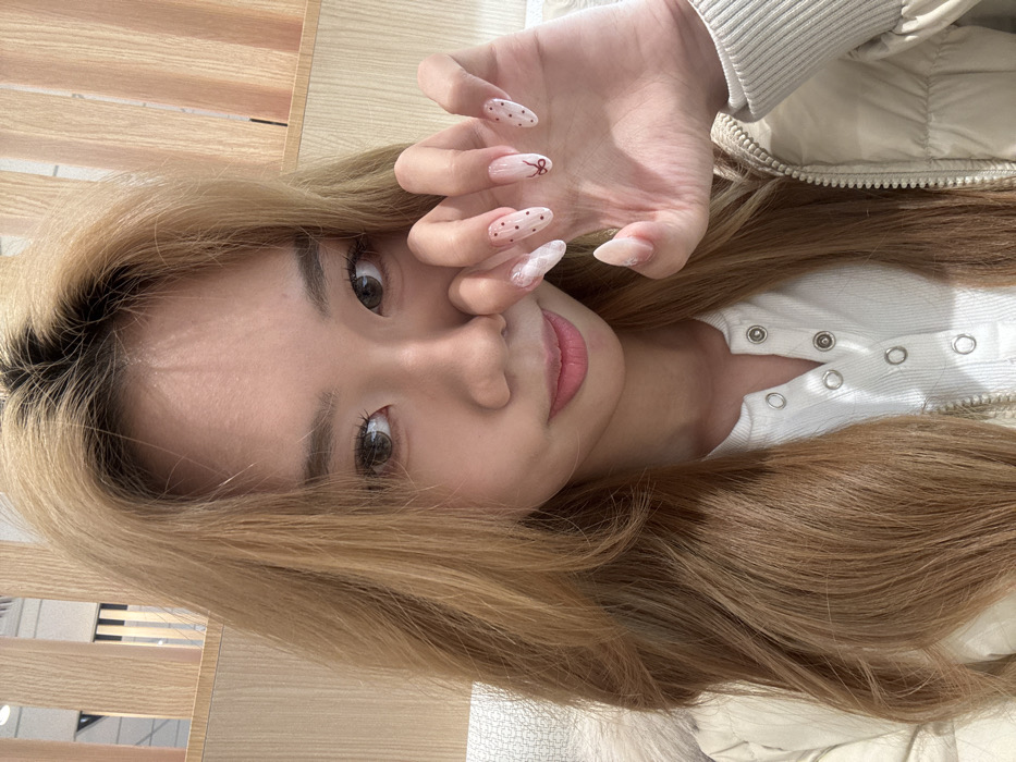 An indoor, chest-up portrait depicts a female subject looking directly at the camera within Yuzhong District, China. She has long, wavy, light brown hair with darker roots, parted centrally. Her eyes are dark brown, augmented with light brown contact lenses and visible makeup. Her lips are subtly pink, curved into a gentle, closed-mouth smile. She wears a white ribbed Henley-style top with silver snap buttons, covered by a light beige puffer jacket with a visible zipper and ribbed cuff on her right wrist. Her right hand is raised, partially obscuring the lower left side of her face. Her nails are long, almond-shaped, and feature a light base with white marble patterns on the thumb and pinky, small red polka dots on the index and middle fingers, and a red bow design on the ring finger. The background is softly out of focus, showing vertical light-colored wooden panels and a textured light object on the far left.