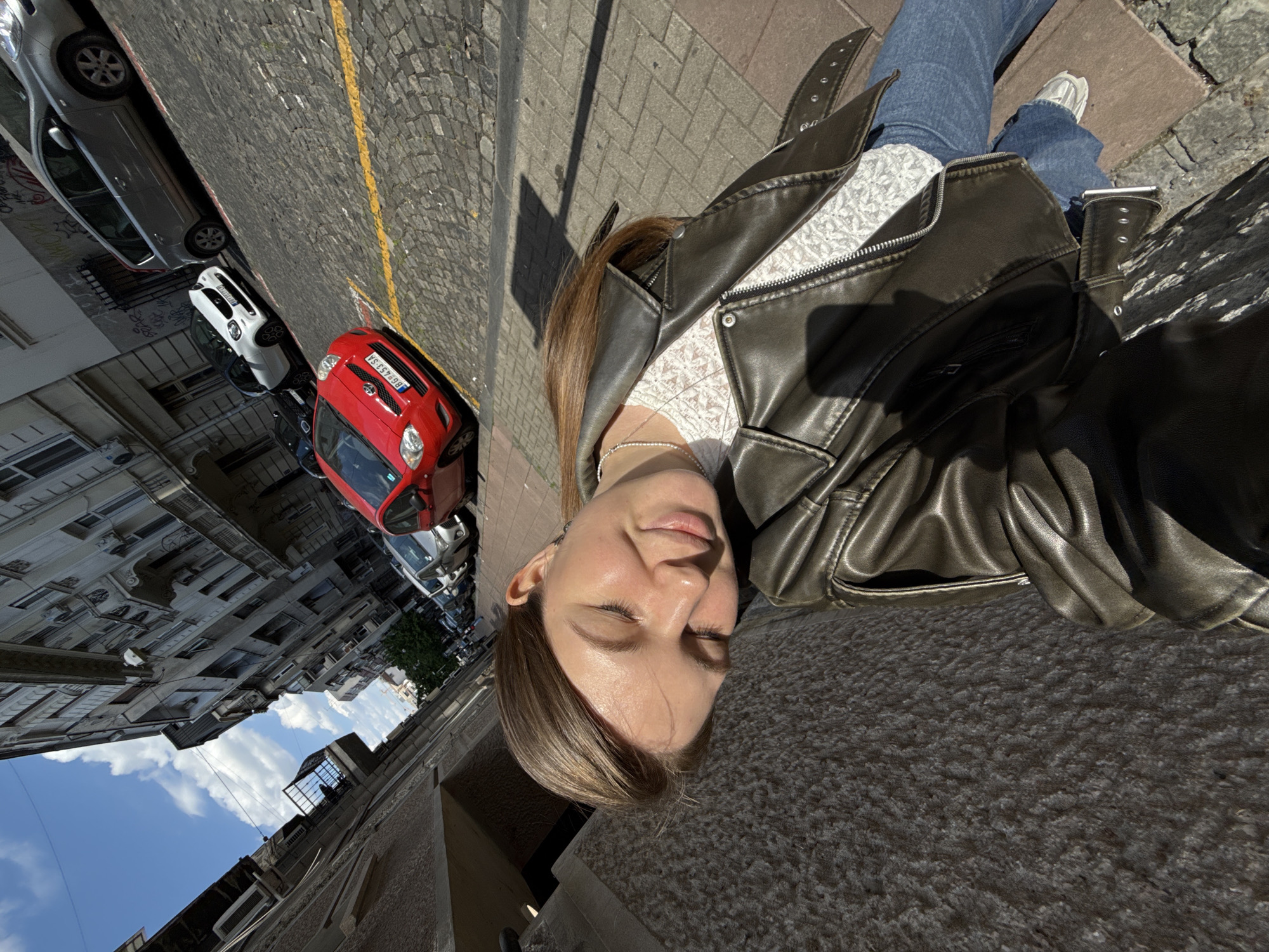 A young woman is captured in a selfie taken outdoors, featuring a street scene and architecture in what appears to be Belgrade, Serbia. The image is taken from a slightly high angle, looking down. The woman, with shoulder-length brown hair, wears a dark brown or olive-toned leather jacket over a white, subtly patterned top, blue jeans, and white sneakers. Her eyes are closed, and she has a soft, relaxed expression, possibly enjoying the sunlight.
She stands on a textured, light grey pavement, with strong shadows cast by the bright sun, indicating a clear, sunny daytime, likely mid-day. To her left, a cobblestone street with a yellow painted line is visible. Several cars are parked along the street. A prominent red compact car, possibly a Toyota Aygo, is closest to the camera, displaying a Serbian license plate "BG 655-SA," where "BG" signifies Belgrade. Behind it, a white car and a silver car are also parked.
Beyond the street, a row of multi-story, older architectural buildings with balconies lines the left side of the frame, some with visible wear and graffiti. Above the buildings, a bright blue sky with scattered white clouds can be seen. The overall scene suggests a vibrant urban environment under pleasant weather conditions.