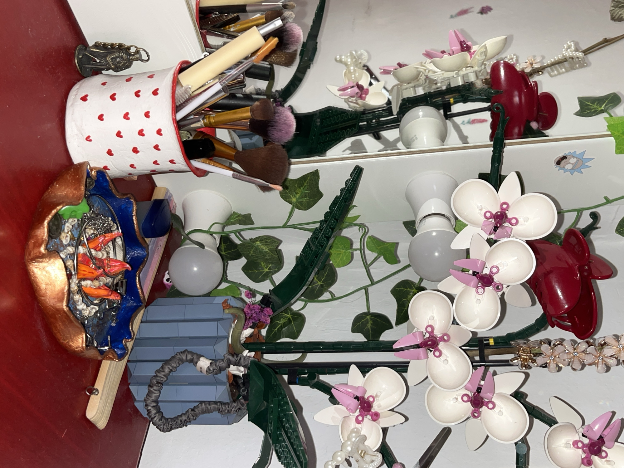 The image, originally rotated 90 degrees clockwise, captures a densely arranged collection of objects on an indoor surface, likely a shelf or desk. The scene is a still life, devoid of any human presence or activity.
On the left, a white ceramic mug, adorned with numerous small red hearts and a red rim, is filled with various makeup brushes. The brushes have different bristle colors, including brown, purple, and beige, and are of varying sizes. To the left of the mug, a small, dark, metallic figurine, possibly a religious or historical figure, stands. Below the mug, a distinctive, multi-colored decorative object, resembling an open, ornate shell or bowl, rests on a light wooden base. Its interior holds small, orange, fish-like elements against a dark blue and black background, while the exterior boasts a coppery, metallic finish.
The central and right areas of the image are dominated by an intricate LEGO botanical set, specifically an orchid arrangement. This structure features multiple white orchid blooms with delicate pink accents, supported by green stems and structural elements that mimic a natural plant. Artificial ivy vines with green leaves are interwoven throughout and behind the LEGO construction, enhancing the botanical theme.
Scattered among the LEGO orchids and vines are several white, frosted, spherical light bulbs, likely modern LED types. A prominent, glossy, dark red hair claw clip is visible among the upper orchids. Below it, a decorative hair accessory, embellished with small, pearl-like beads and clear, flower-shaped accents, adds another touch.
A small sticker depicting the head of Rick Sanchez from the animated series "Rick and Morty" is affixed to the white background surface. Near the bottom-left of the LEGO arrangement, a grey corrugated tube, possibly a cable management sleeve, is seen adjacent to a section of grey, stepped LEGO bricks. The background consists of a white wall or shelf, with a subtle reflection suggesting a mirror or glossy surface above the main display. The lighting appears to be indoor and artificial.