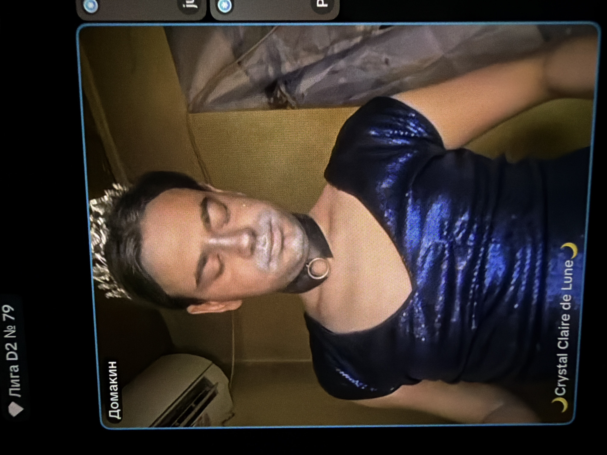 This media file is a rotated image, appearing to be a photograph of a digital screen, showing a person in an indoor setting. The subject is a young adult with dark hair, wearing a silver crown-like headpiece and a shiny, dark blue, possibly sequined, short-sleeved top or dress. They also wear a black choker with a silver O-ring. A white substance, possibly makeup or powder, is visible on their face, particularly around the mouth and cheeks. Their eyes are closed or mostly closed, and the expression is neutral.
The background features a muted yellow or light brown wall. In the lower-left background, a white electronic device, possibly an air conditioning unit, is visible. The upper background shows a dark, indistinct fabric or object. The person appears to be posing for a photo, possibly for a costume event or performance.
Visible text overlays on the image, indicating it's a screenshot or photo of an interface. On the left side, rotated, are "Лига D2 № 79" and "Домакин". On the right side, also rotated, is "☽ Crystal Claire de Lune". Partially visible UI elements, including "ju" and two circular icons, are present at the top. The image itself provides no direct visual cues to the specified location of Sofia, Bulgaria.
