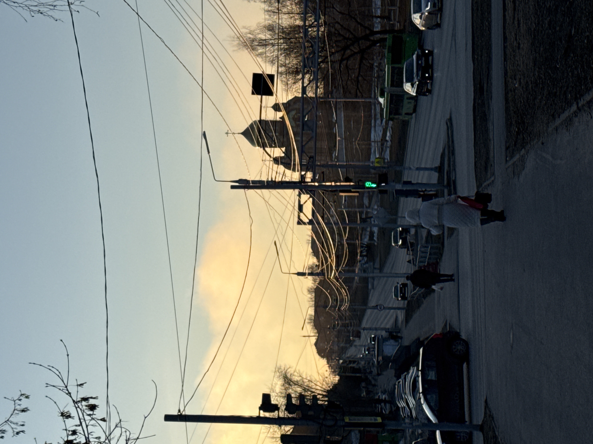 The image depicts a street in Berezniki, Russia, at dusk. The sky is a gradient of blue and orange. Overhead power lines crisscross the view. A church with a prominent dome is visible amidst the buildings. A traffic light shows a green light. People walk along the sidewalk on the right. Cars are parked along the side of the road.
