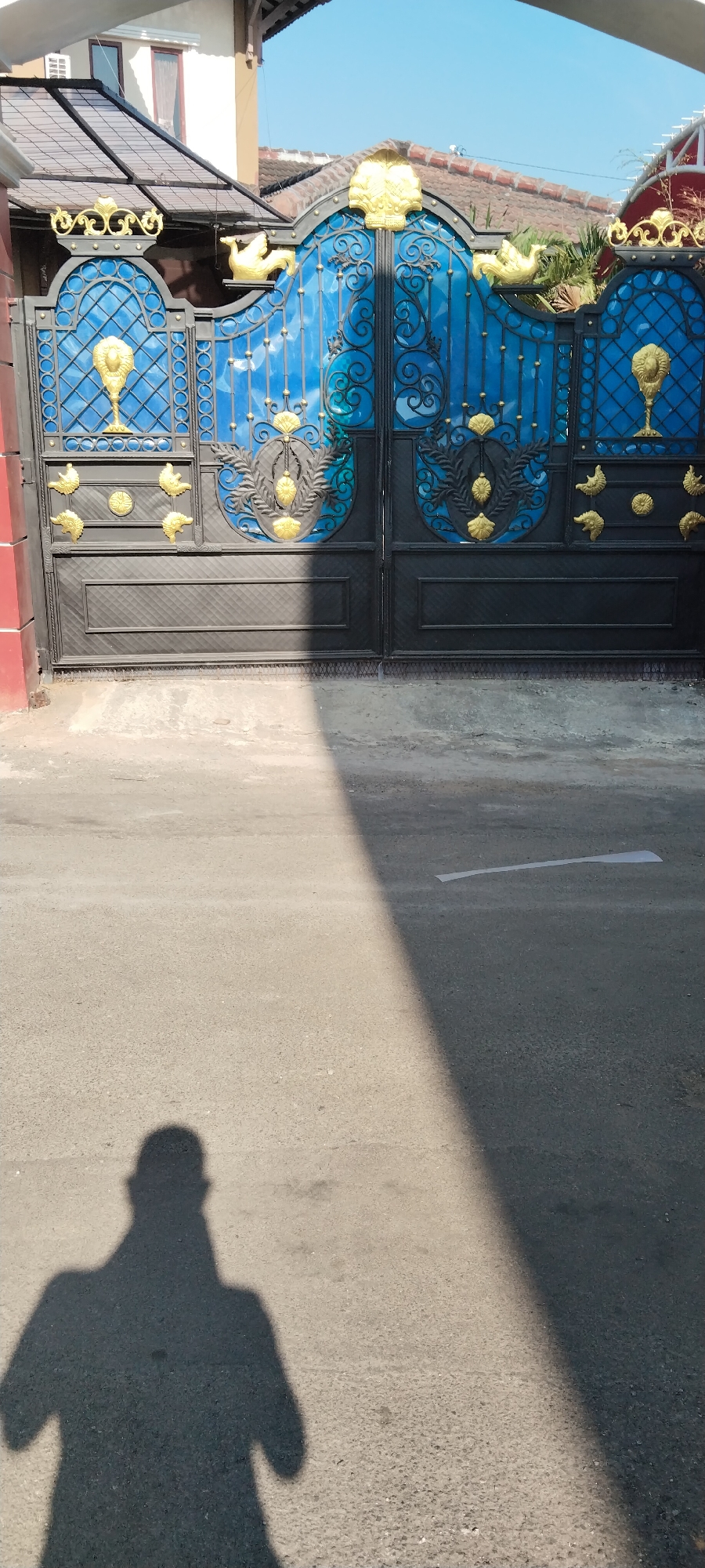 The image shows a black metal gate with ornate gold embellishments, including what appear to be chalices, shells, and birds. The gate has blue glass panels behind the metalwork. In front of the gate is an asphalt road, and a shadow of a person is cast across the road, indicating it is daytime. The location is identified as Ungaran, Indonesia, suggesting this is likely a residential gate. The scene is static and captured from a slightly elevated perspective looking down at the gate and road.