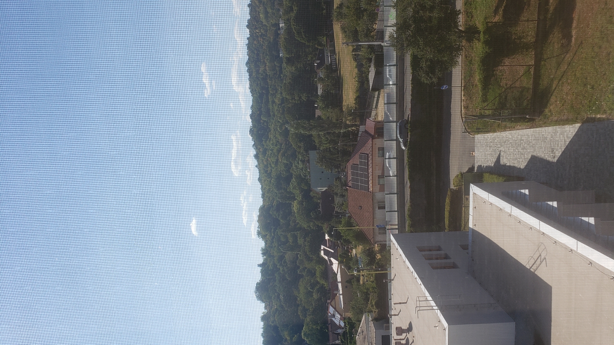 This is a photograph taken through a window screen, looking out over a suburban area. In the foreground, the roof and side of a modern, light-colored building are visible. Beyond this building, a road runs horizontally across the frame, with a dark-colored car driving on it. A transparent barrier separates the road from a row of houses with red-tiled roofs, some of which have solar panels. The houses are nestled amongst lush green trees that form a dense forest covering a rolling hill in the background. The sky is a clear blue with a few small white clouds. The scene appears to be a residential area on the outskirts of a city, likely in Slovakia, given the mention of Prešov. The overall impression is one of a sunny, peaceful day in a verdant landscape.