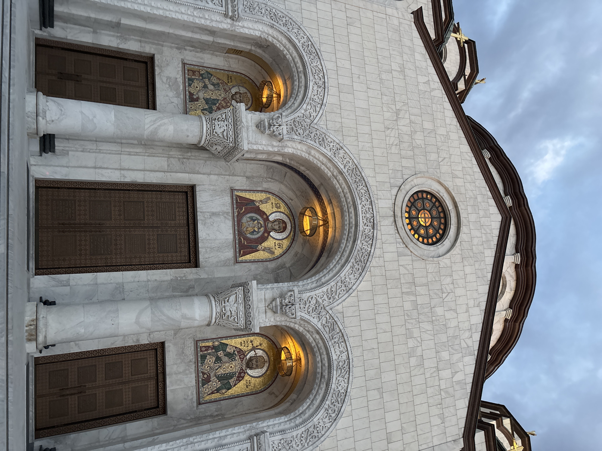 The image shows the facade of the Church of Saint Sava in Belgrade, Serbia. The church is constructed of white marble, with a large central dome and several smaller domes. The facade features three arched entrances, each adorned with intricate carvings. Above the central arch is a circular stained-glass window. Flanking the entrances are mosaics depicting religious figures. The doors themselves are made of bronze and feature decorative patterns. The sky above is cloudy and grey.