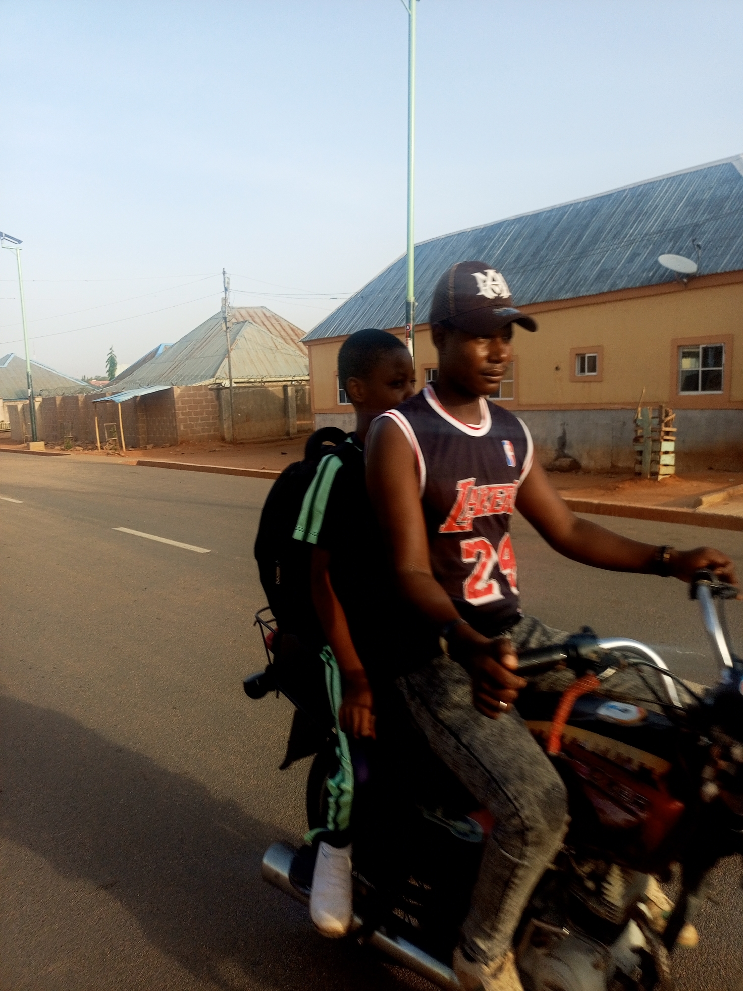 A man is riding a motorcycle down a street in Unknown, Unknown. He is wearing a black cap, a black and white Lakers jersey with the number 24 on it, and grey pants. A young boy is sitting behind him with a black backpack with green stripes. They are on a paved road lined with buildings. There is a street light on the side of the road.
