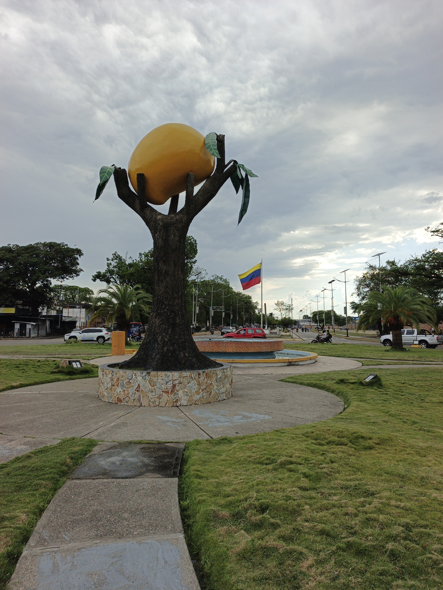 The image depicts a public space in San Carlos, Venezuela. Dominating the scene is a large sculpture of a mango tree with a single, oversized, ripe mango fruit atop its branches. The tree trunk is painted dark brown and sits on a circular stone base.
In the background is a Venezuelan flag flying near a roadway. Traffic, including cars and motorcycles, is visible. Other palm trees and greenery are in the vicinity.
The sky is overcast with gray clouds, suggesting potentially humid weather. The ground is covered in well-maintained grass. A concrete walkway leads up to the mango sculpture.