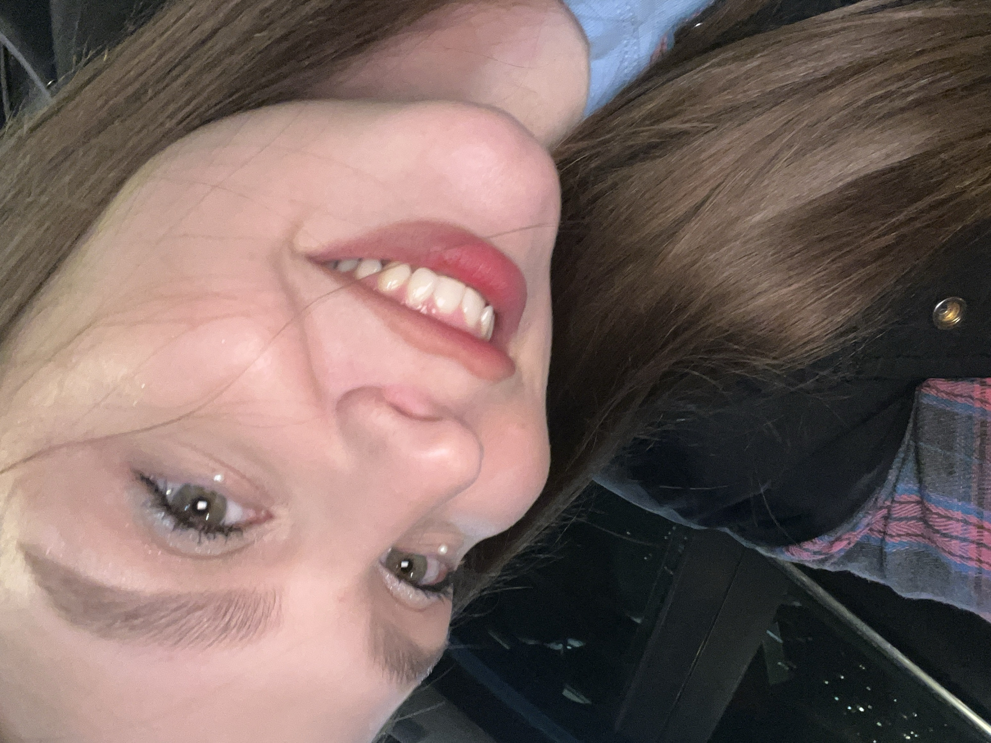 The image features a close-up, rotated view of a young woman's face and upper body, appearing to be a selfie or a casual portrait. Her head is significantly tilted to the left, with her hair falling to the right side of the frame. She has long, straight brown hair.
Her face shows a clear, bright left eye with a green/hazel iris and visible eyeliner and mascara. Her left eyebrow is well-defined. Her nose and mouth are also prominent, with a wide, open-mouthed smile revealing several white teeth and red lipstick on her lips. Her skin appears fair.
She is wearing multiple layers of clothing. A light blue fabric is visible near her neck and shoulder. Below that, a darker fabric, possibly a shirt or jacket, is present. A plaid pattern in shades of pink, blue, and black is visible on what appears to be a flannel shirt or similar garment worn over the darker layer. A metal button or snap is visible on the dark fabric near her hair.
The background is dark and indistinct, suggesting an indoor setting or an outdoor nighttime environment. There are some blurry light reflections or distant lights in the deep background, along with dark, geometric shapes that could be parts of furniture, architecture, or a window frame. A thin, metallic-looking bar or railing is also visible in the lower right background.
The woman's smiling expression conveys a sense of happiness or contentment. There is no visible text in the image. Given the location context of Kudrovo, Russia, the scene is likely indoors within that area.