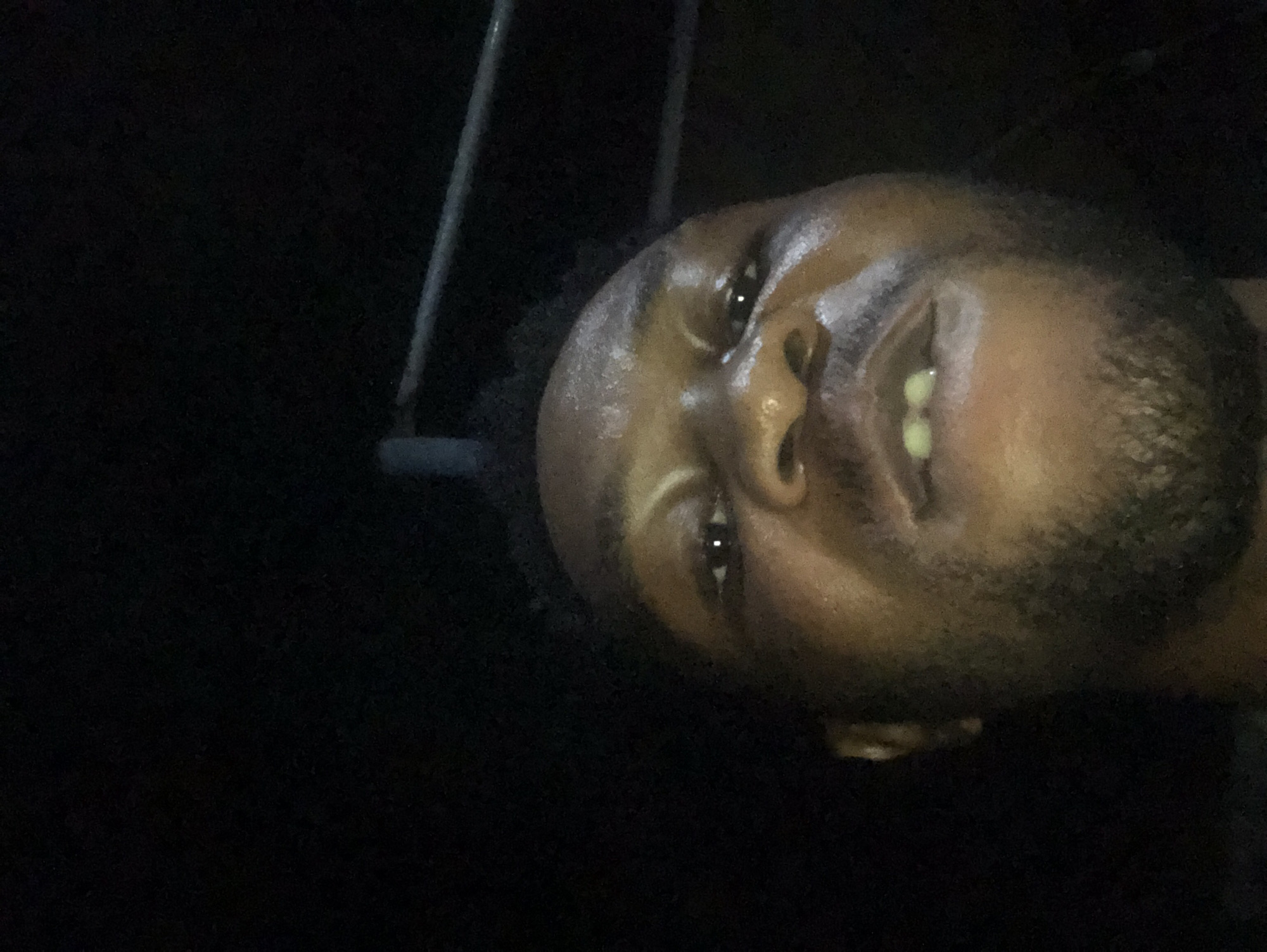 Here is a description of the image:
A selfie of a dark-skinned man with a short beard. His face is angled, and he is looking at the camera with a slight smile. The lighting is low, and the background is mostly dark except for some light elements. The location is Unknown, Unknown.
