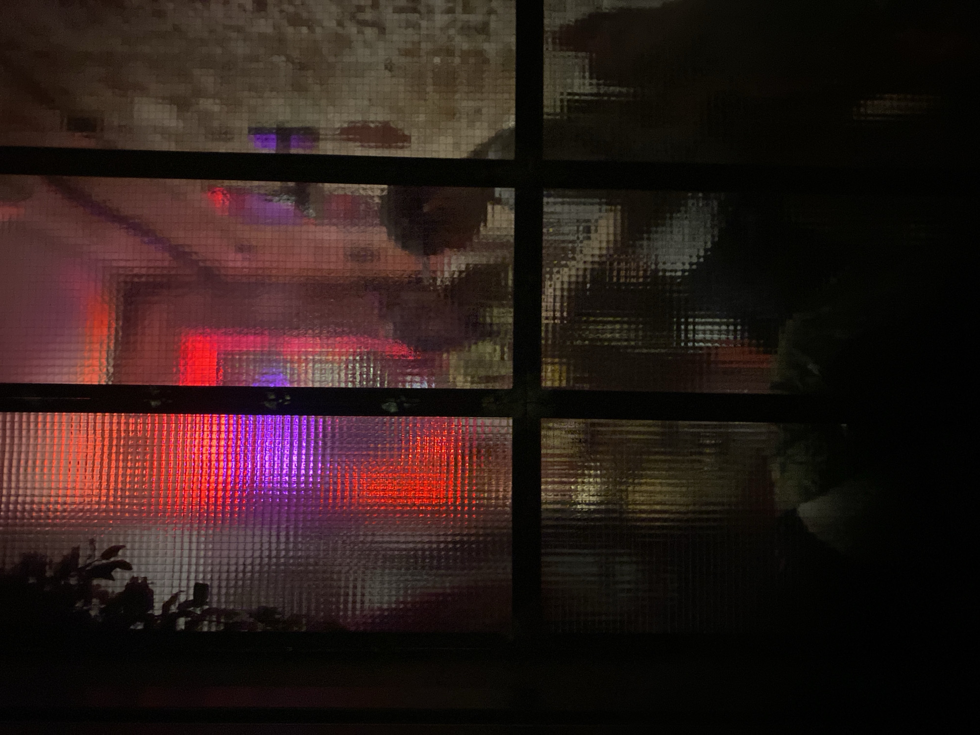 The image shows a view through a window with a grid pattern. Outside, there are blurry lights in red, orange and purple. The building exterior appears to be beige with a tile-like pattern. Some vegetation is visible at the bottom of the window. The right side of the image is dark, obscuring details. The location is presumed to be Moscow, Russia. There are indications of people's shadows and movement outside.