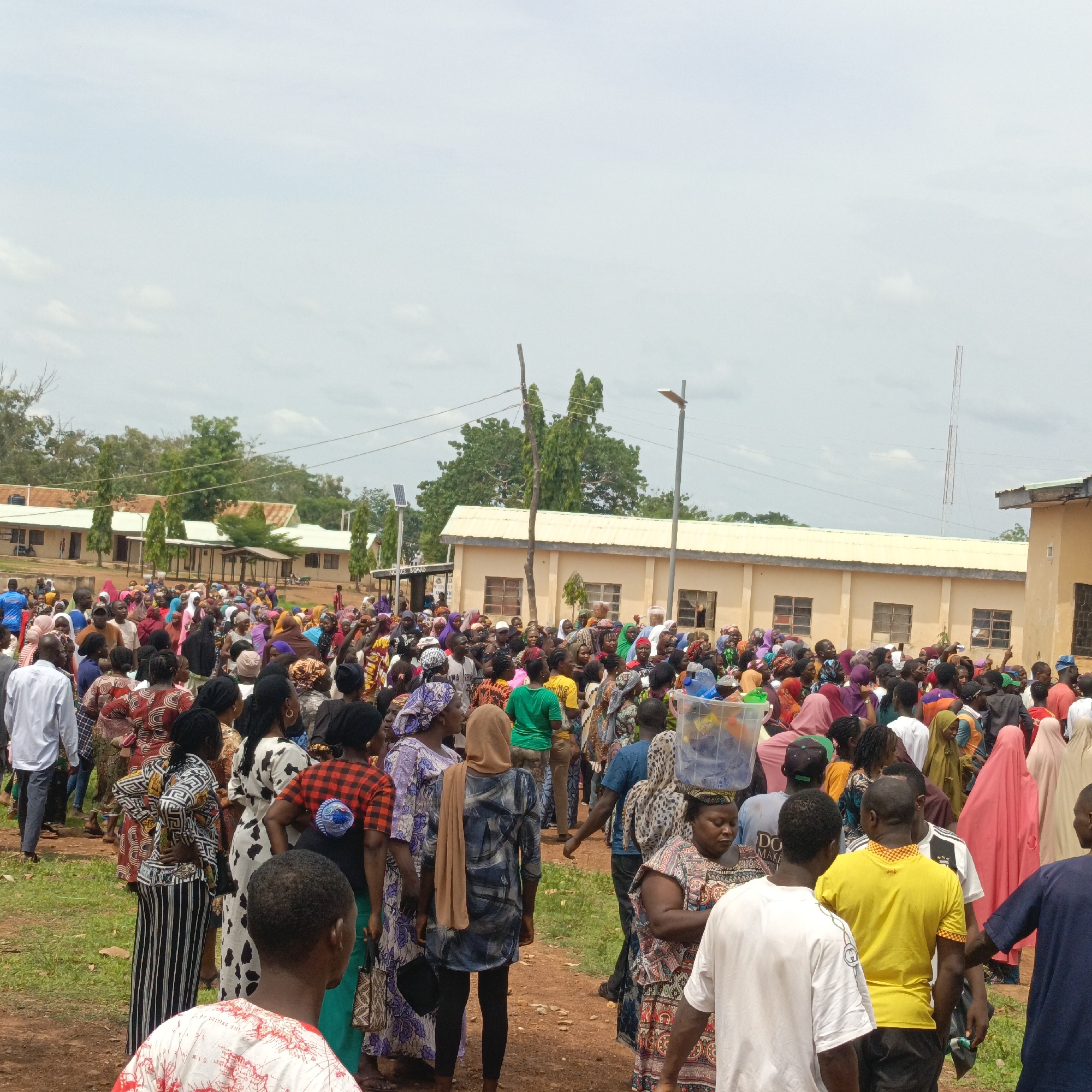 The image depicts a large crowd of people congregated in an open area in what appears to be Akwanga, Nigeria. The majority of individuals are dressed in colorful traditional attire, with many women wearing headscarves. The crowd is dense, suggesting a gathering or event of some significance. Buildings with a light-colored exterior and low rooftops are visible in the background, possibly indicating a public or community space. The ground is a mixture of dirt and grass. One woman carries a clear container on her head. Several men are wearing western-style clothing. In the upper portion of the image, the sky is overcast.