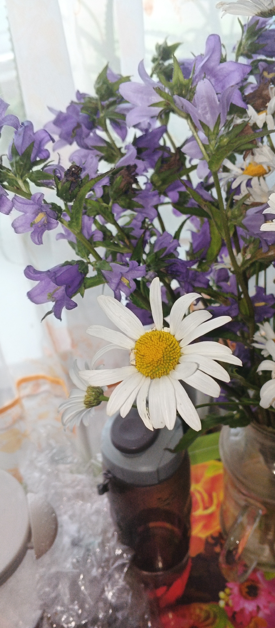 A close-up, slightly angled shot focuses on a bouquet of wildflowers, primarily purple bellflowers and white daisies, placed in a vase. The flowers are in the foreground, with the purple bellflowers forming a dense backdrop behind the prominent white daisies. One daisy, in sharp focus, displays a bright yellow center and white petals, some of which appear slightly wilted. Behind the flowers, a sheer white curtain is visible, suggesting an indoor setting near a window. To the right and bottom of the frame, portions of various objects are discernible: a dark brown plastic bottle with a gray lid, possibly a water bottle, and a glass vase containing more flowers. The surface beneath the vases is decorated with a vibrant, colorful floral pattern. A translucent, crinkled material, perhaps plastic packaging, is present on the left side of the image. The scene appears to be a still life composition, possibly arranged in a home or a studio environment in the city of Podgornoye, Russia.