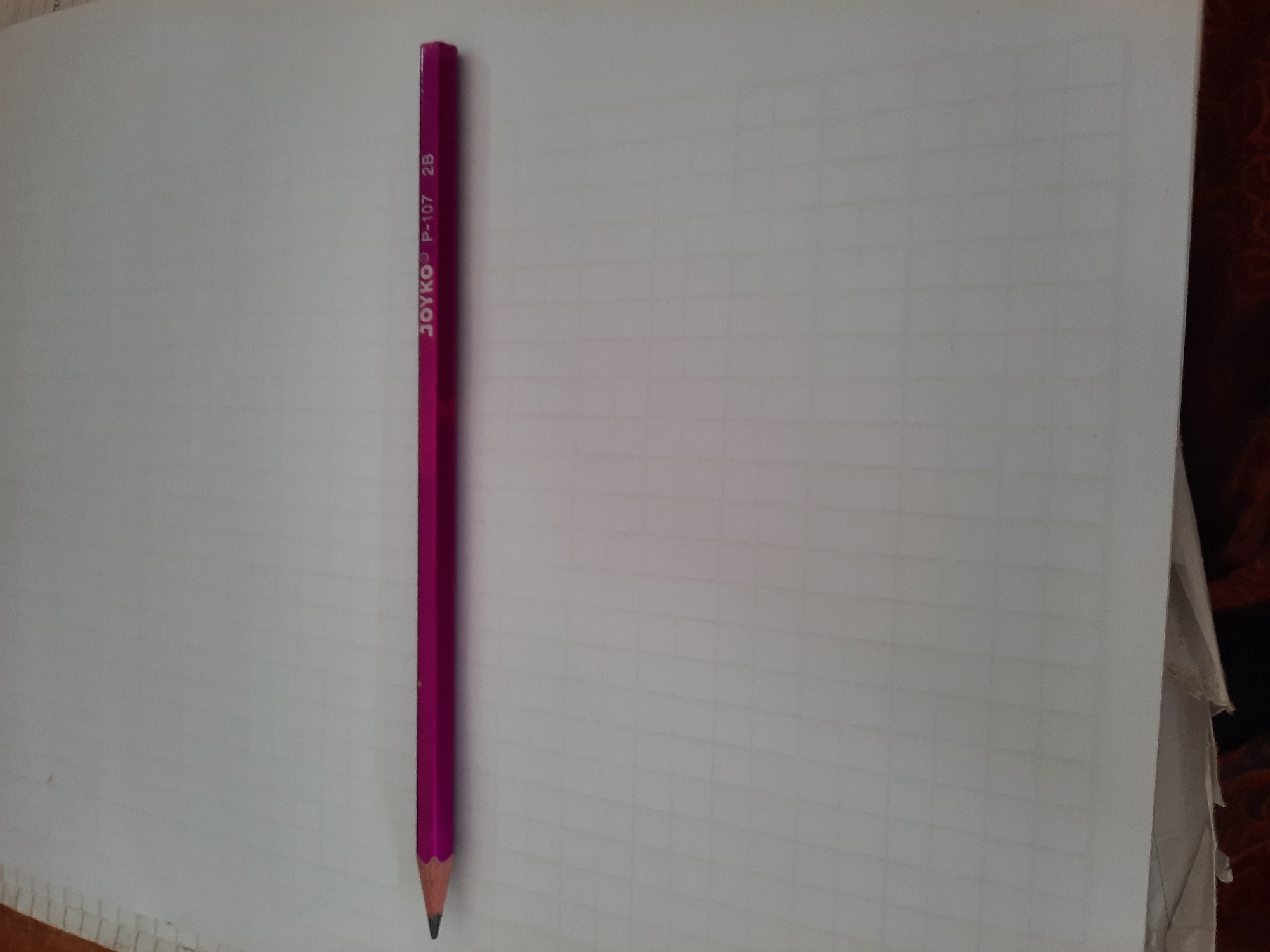 An image shows a pink pencil with the brand name "JOYKO P-107 2B" printed on its side, lying horizontally on a piece of graph paper. The pencil is positioned in the center of the frame, with its sharpened tip pointing to the left. The graph paper has faint gray lines forming a grid. The background is plain white. The image does not provide any information about the location in Lhokseumawe, Indonesia, or any people or actions.
