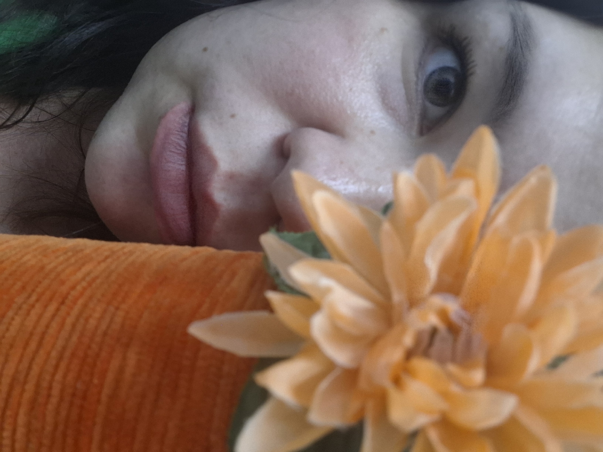 A close-up, shallow depth-of-field shot features a person partially visible, an orange ridged fabric, and a blurred artificial flower. The person's face is seen from the nose to the left side of the mouth and one wide-open eye looking towards the viewer. The skin appears fair with some freckles and a slight redness on the cheek. Her lips are a soft pink, slightly parted, with a defined texture. The eye, with a dark iris, is framed by long, dark eyelashes and a visible eyebrow. Dark hair, with a hint of green, is visible in the upper left corner, suggesting it belongs to the person.
The person appears to be lying down, with her head resting on an orange, ribbed fabric surface, possibly a pillow or blanket, which occupies the lower left portion of the frame. In the foreground, a large, light orange or peach-colored artificial flower is prominently placed, largely out of focus, creating a soft, blurred effect. Its numerous petals are distinguishable despite the blur.
The image has a soft, intimate feel, focusing on facial details and textures. The lighting is diffused, highlighting the contours of the face and the fabric. There is no visible text. The setting appears to be indoors.