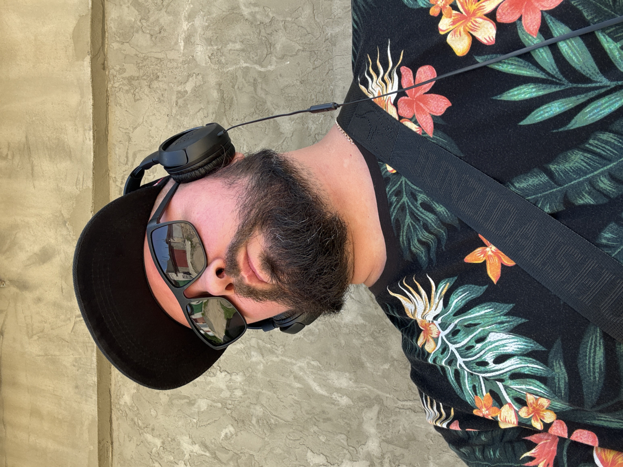 A close-up, low-angle shot captures a man wearing a black baseball cap, dark sunglasses, and over-ear headphones. He has a beard and is wearing a black t-shirt with a vibrant floral and leaf pattern. A black strap, likely from a bag, is visible diagonally across his chest. The background is a textured, light-colored wall. The scene is set outdoors, suggested by the reflection of buildings and a street in the man's sunglasses and the bright sunlight. The man is looking upwards and slightly to his left, with his head tilted. The image does not provide any specific information or context to indicate a location in Orsha, Belarus.