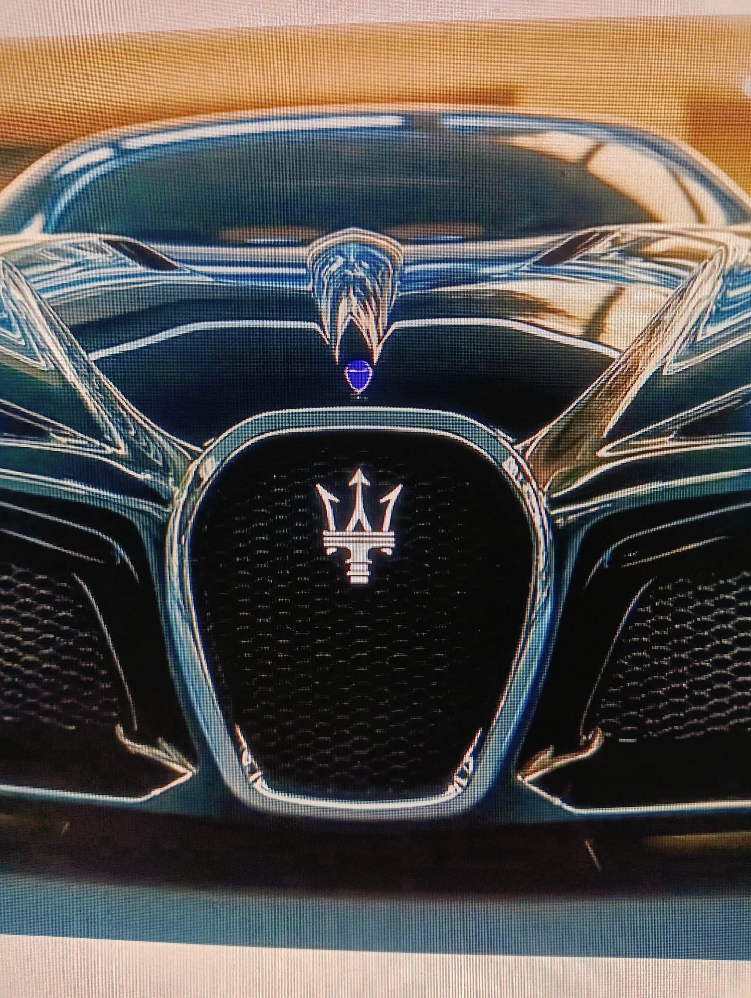 The image displays a close-up, frontal view of a dark-colored luxury car's grille and emblem. The car's surface is highly polished, reflecting the surrounding environment which appears to be outdoors with a muted, tan-colored backdrop, possibly suggesting a wall or building. The dominant feature is the prominent chrome grille, characterized by a textured mesh pattern. Centered on the grille is the car's emblem, a stylized trident, also in a metallic finish. Above the grille, on the hood, is a smaller, blue emblem with a chrome outline. The car's headlights and parts of its hood are visible, designed with sharp, angular lines, contributing to its aggressive aesthetic. There are no people or any other distinguishing features that would suggest a specific location within the city of Sluck, Belarus, or any other urban environment. The image is a static, focused shot of the vehicle's front fascia.