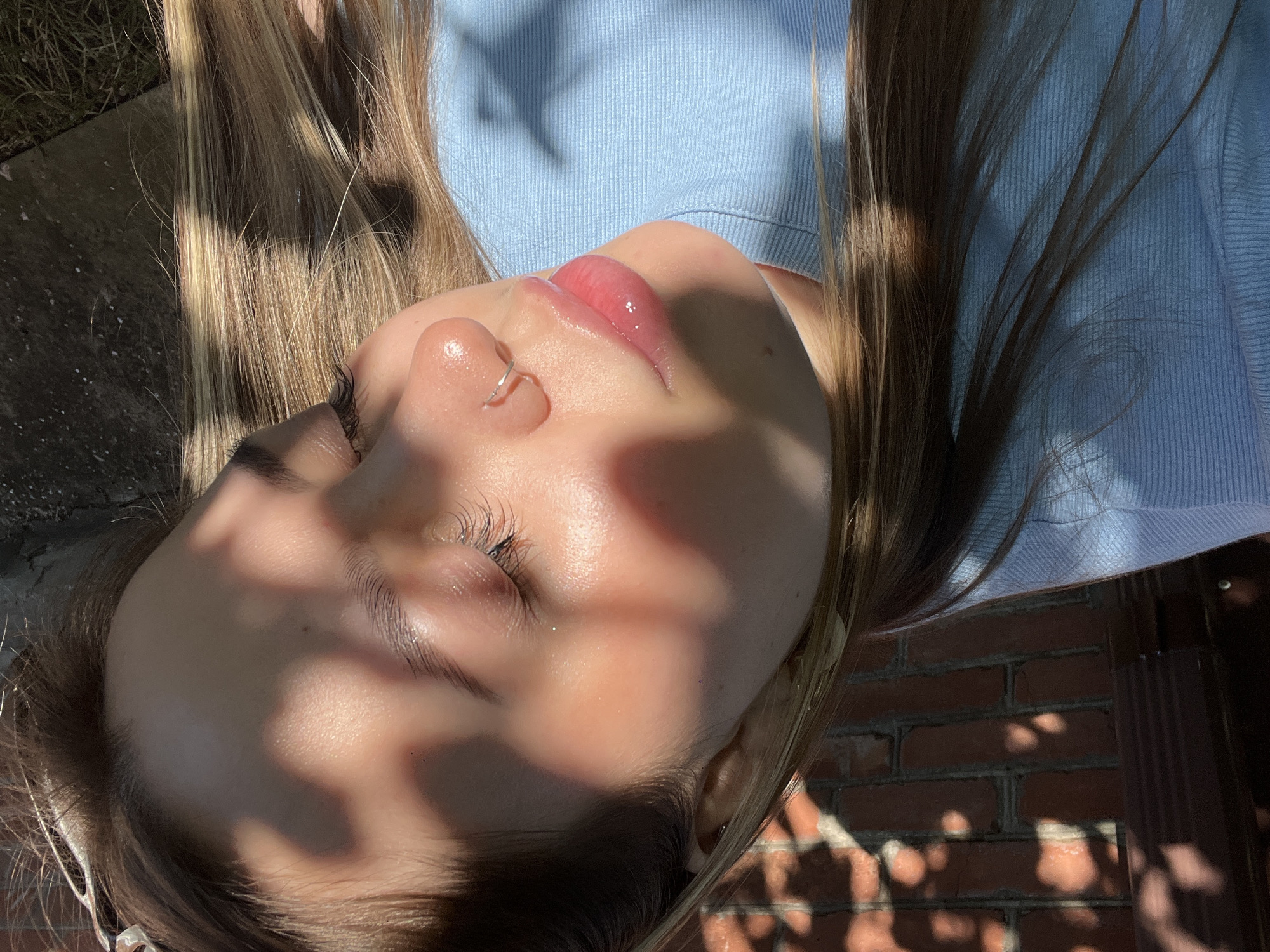 The image captures a close-up, tilted view of a person, likely female, resting outdoors on a sunny day. Her eyes are closed, highlighting long dark eyelashes, and her lips are glossy with a pink tint. A small, shiny nose piercing adorns her right nostril. Long, light brown hair with blonde highlights frames her face and spreads over a light blue, ribbed shirt. Distinct shadows from unseen foliage dapple her skin and hair, creating a play of light and shade. In the background, sections of a reddish-brown brick wall and concrete are visible, indicating an outdoor setting. The strong, direct sunlight suggests a bright, clear day. The scene evokes a feeling of calm and warmth. No text is visible in the image.