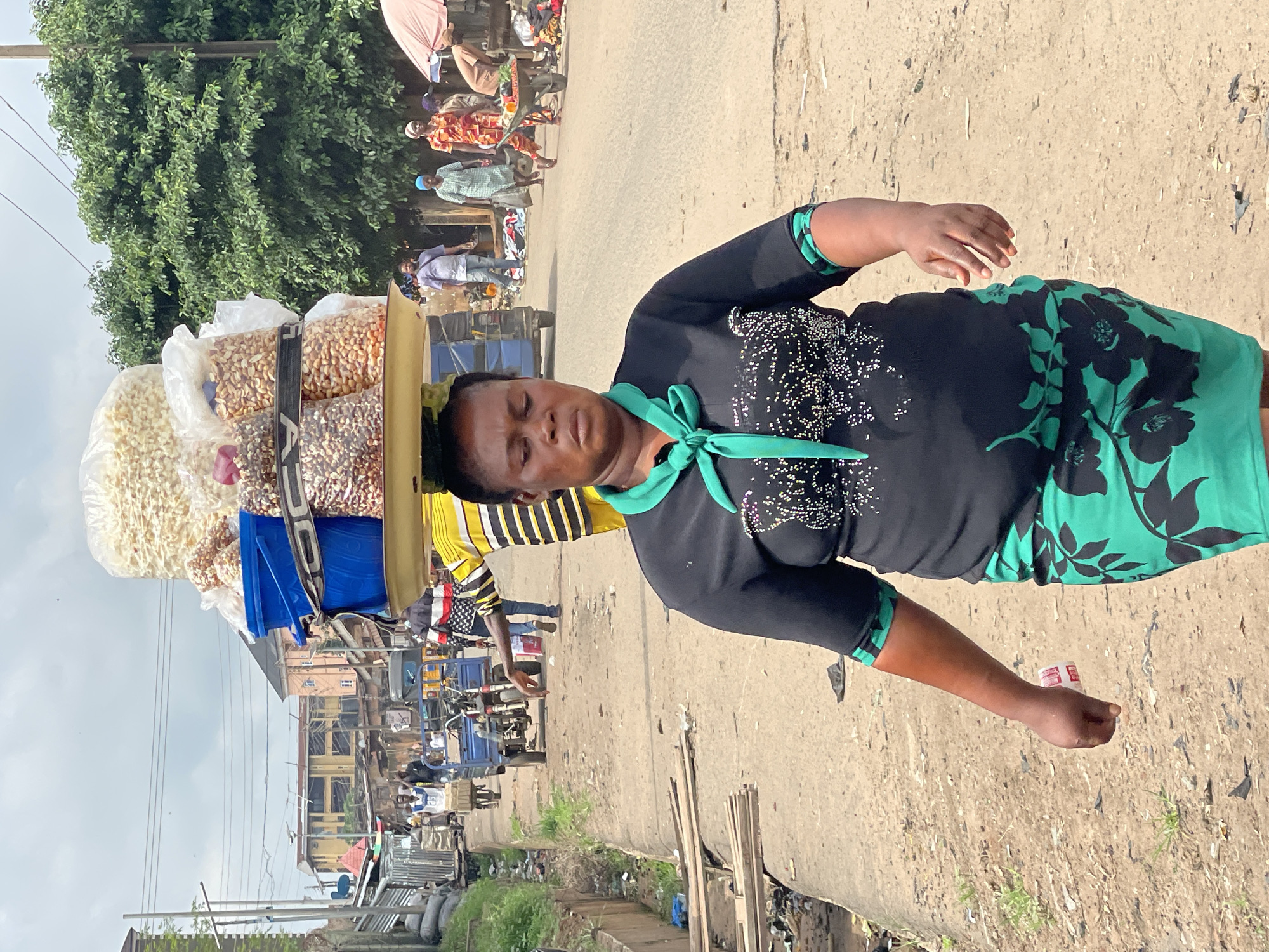 A woman walks along a dirt road, carrying a large tray of goods balanced on her head. The tray contains multiple bags of what appears to be nuts and popcorn, along with a blue bucket. She is wearing a black short-sleeved shirt with a green collar and a green and black floral skirt. Her expression is neutral as she walks towards the viewer. In the background, several other people and market stalls are visible, along with a few vehicles, including a tricycle. The environment appears to be a busy outdoor market or street scene in a city.