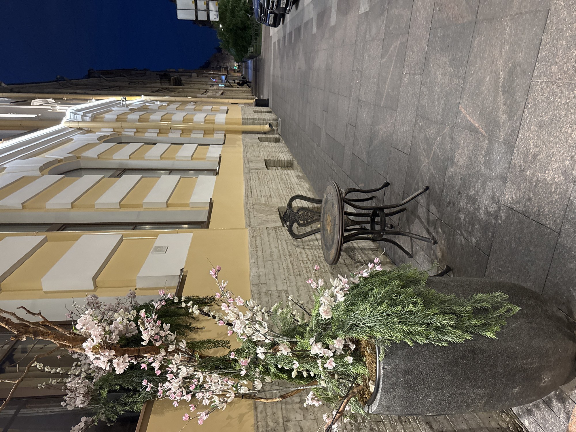 The image depicts a sidewalk scene at night on Nevsky Prospekt in Saint Petersburg, Russia. On the left, a large, dark gray stone planter holds a decorative tree with delicate pink and white blossoms and trailing green foliage. Adjacent to the planter, a small, ornate, dark metal table with a single matching chair is set on the stone-tiled sidewalk. The building to the right of the planter has a pale yellow facade with white decorative trim and arched windows, illuminated by interior lights. The sidewalk extends into the distance, lined with parked cars on the right. The sky is a deep, dark blue, indicating it is nighttime. A single pedestrian is visible in the distance, walking away from the camera. The scene appears to be an outdoor seating area for a establishment.