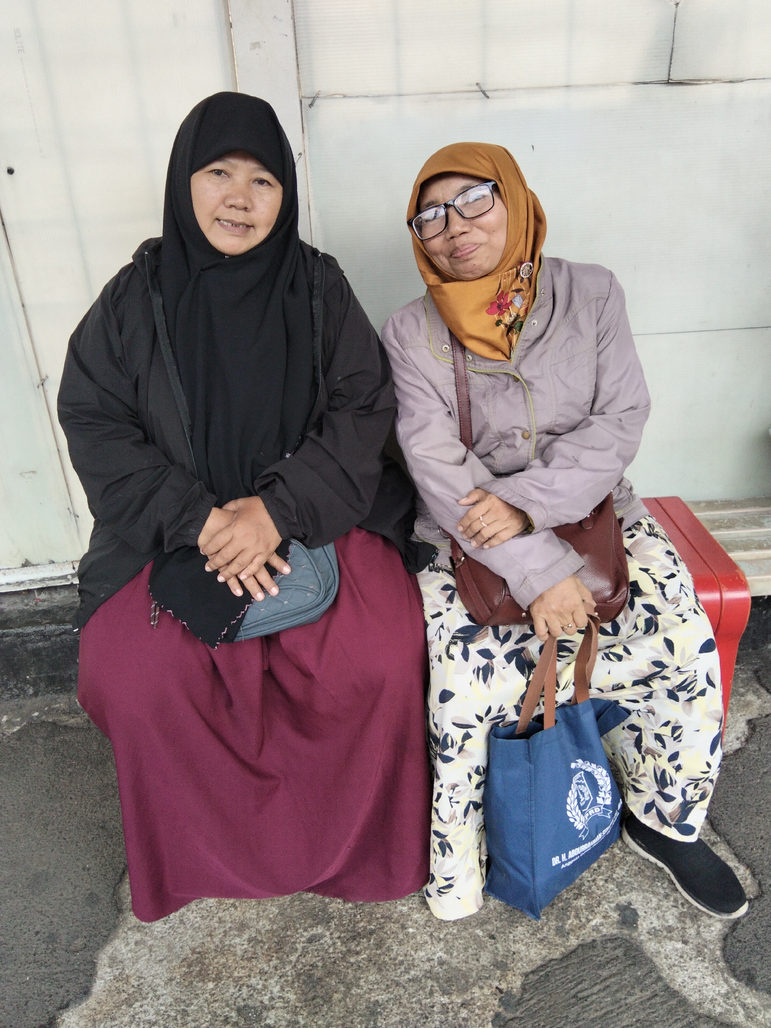 Two women are seated side-by-side on a red bench, possibly outdoors in South Jakarta, Indonesia, with a light-colored, textured wall behind them. The woman on the left wears a black hijab and a dark jacket over a maroon skirt. Her hands are clasped in her lap, resting on a small, grey quilted purse. The woman on the right wears a mustard-colored hijab, glasses, and a light purple jacket over a white and floral-patterned skirt. She has a brown leather purse slung over her shoulder and holds the straps of a blue canvas tote bag in her hands. The tote bag has white text and an emblem on it. The ground in front of them is paved with a rough, gray material, appearing to be a sidewalk or pathway. The overall impression is one of casual waiting or resting.