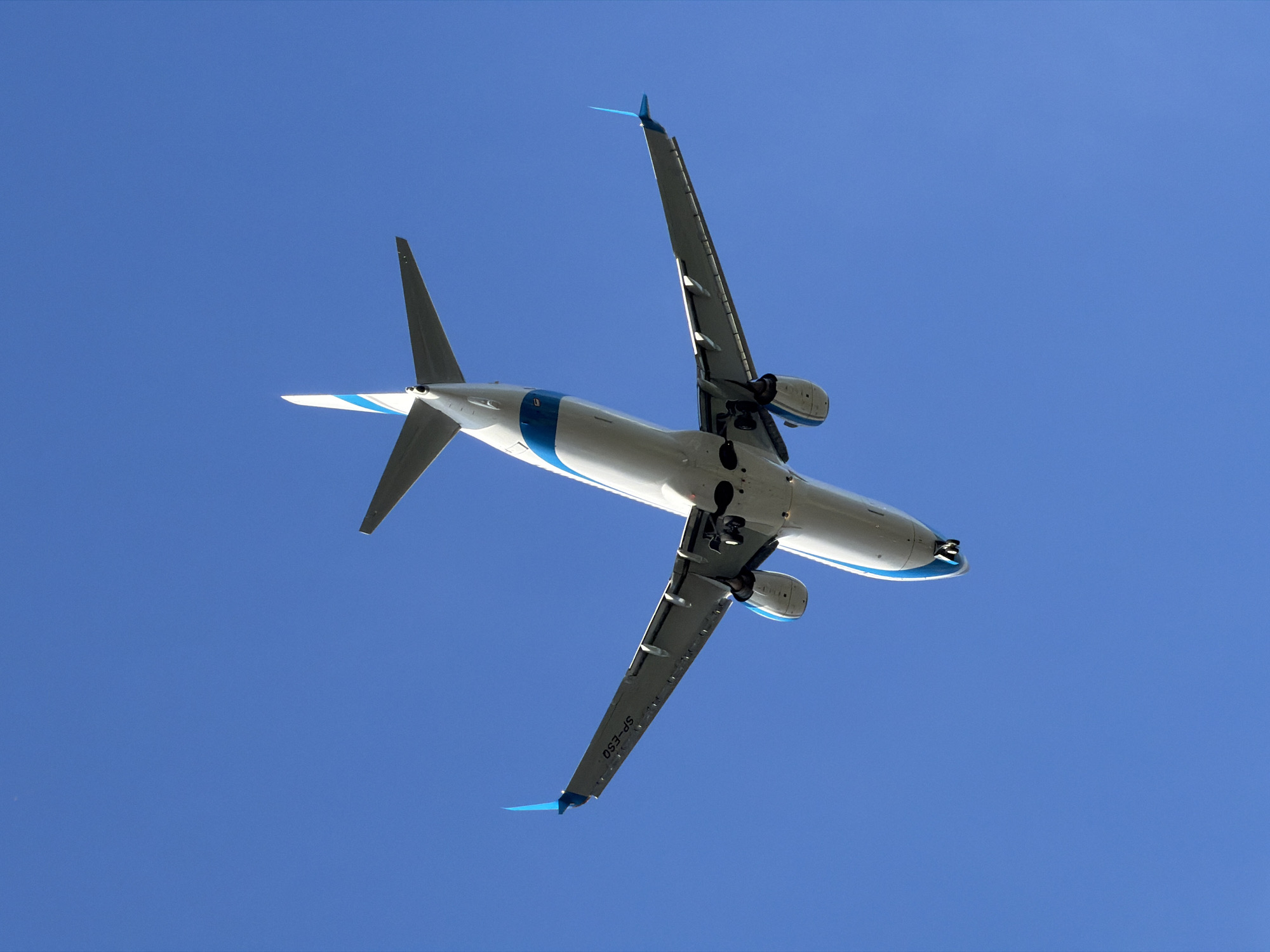 An Airbus A320-200 aircraft, registered as SP-ESO, is captured in an underside view against a clear blue sky. The aircraft, predominantly white with blue accents on its tail fin and wingtips, is in flight, with its landing gear extended. The image focuses on the plane's fuselage, wings, engines, and tail. The writing "SP-ESO" is visible on the underside of the left wing. The angle suggests the aircraft is either ascending or descending. The specific location in Burgas, Bulgaria, is not discernable from the image itself.