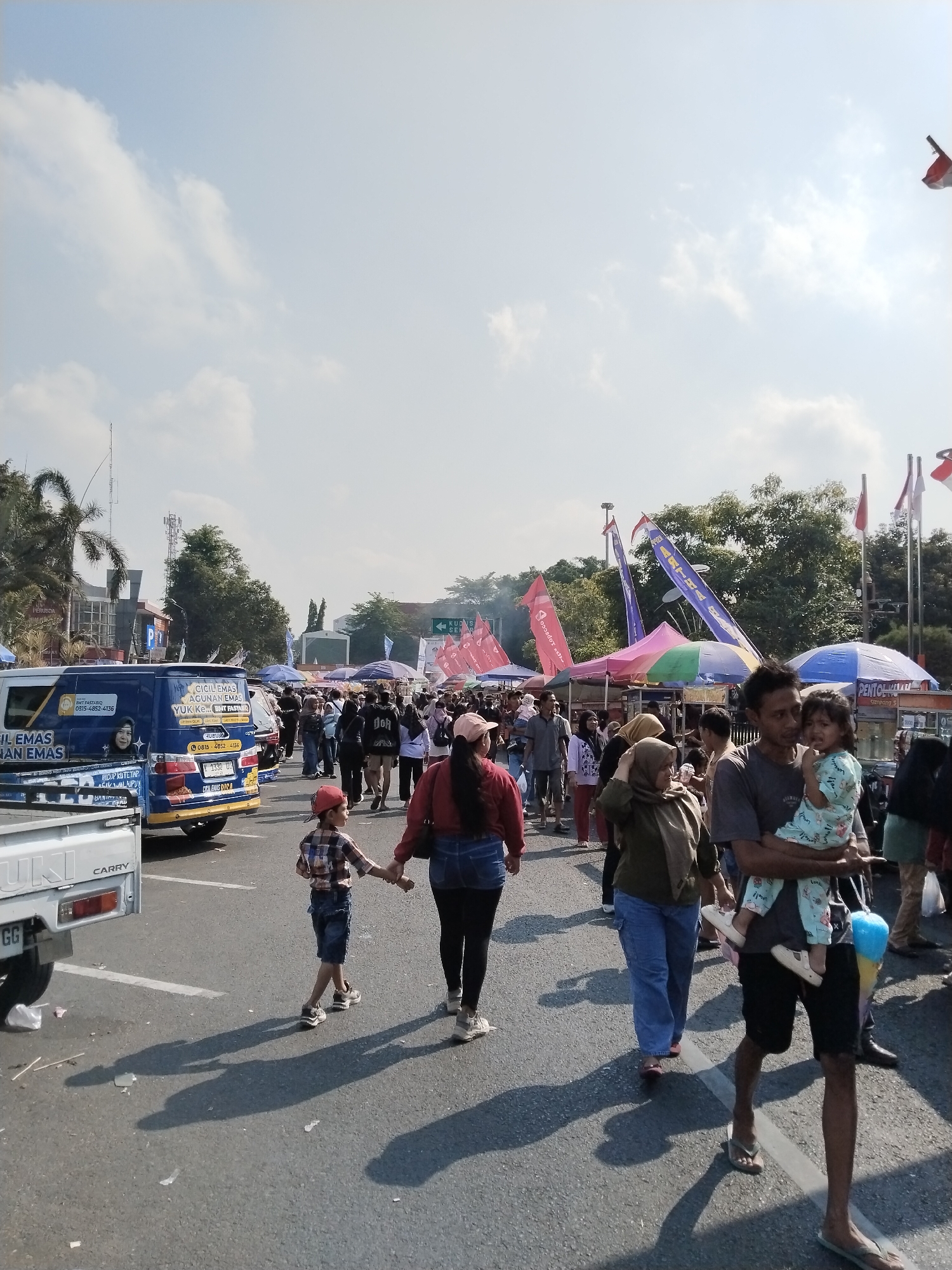 The image depicts a street scene in Kecamatan Pati, Indonesia, likely during a daytime event or market. A crowd of people are walking along a paved road lined with food stalls and vendors under colorful awnings and banners. In the foreground, a young boy in a plaid shirt and red cap walks hand-in-hand with a woman in a red jacket and jeans. To the right, a man holds a baby wearing a light blue patterned outfit. A blue van with advertising is parked on the left side of the road, and a white pickup truck is partially visible. The sky is bright with scattered clouds, and sunlight casts long shadows on the pavement. Red and white Indonesian flags are visible on poles in the background.