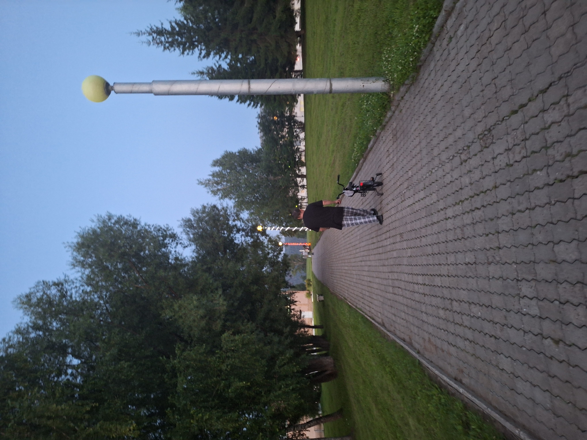 A person is walking along a paved walkway in a park-like setting. The person is wearing a black t-shirt and checkered pants. They are pushing a small black bicycle. The walkway is made of interlocking grey pavers and is lined with grass on one side and trees on the other. There are several trees of varying sizes, some appearing to be deciduous and others coniferous. In the background, there are buildings and more trees. A tall lamppost with a yellow globe on top is visible to the right of the walkway. The sky is a clear, pale blue, suggesting it is either early morning or late evening. The scene appears to be in a residential area or a park in the city of Atamanovka, Russia.