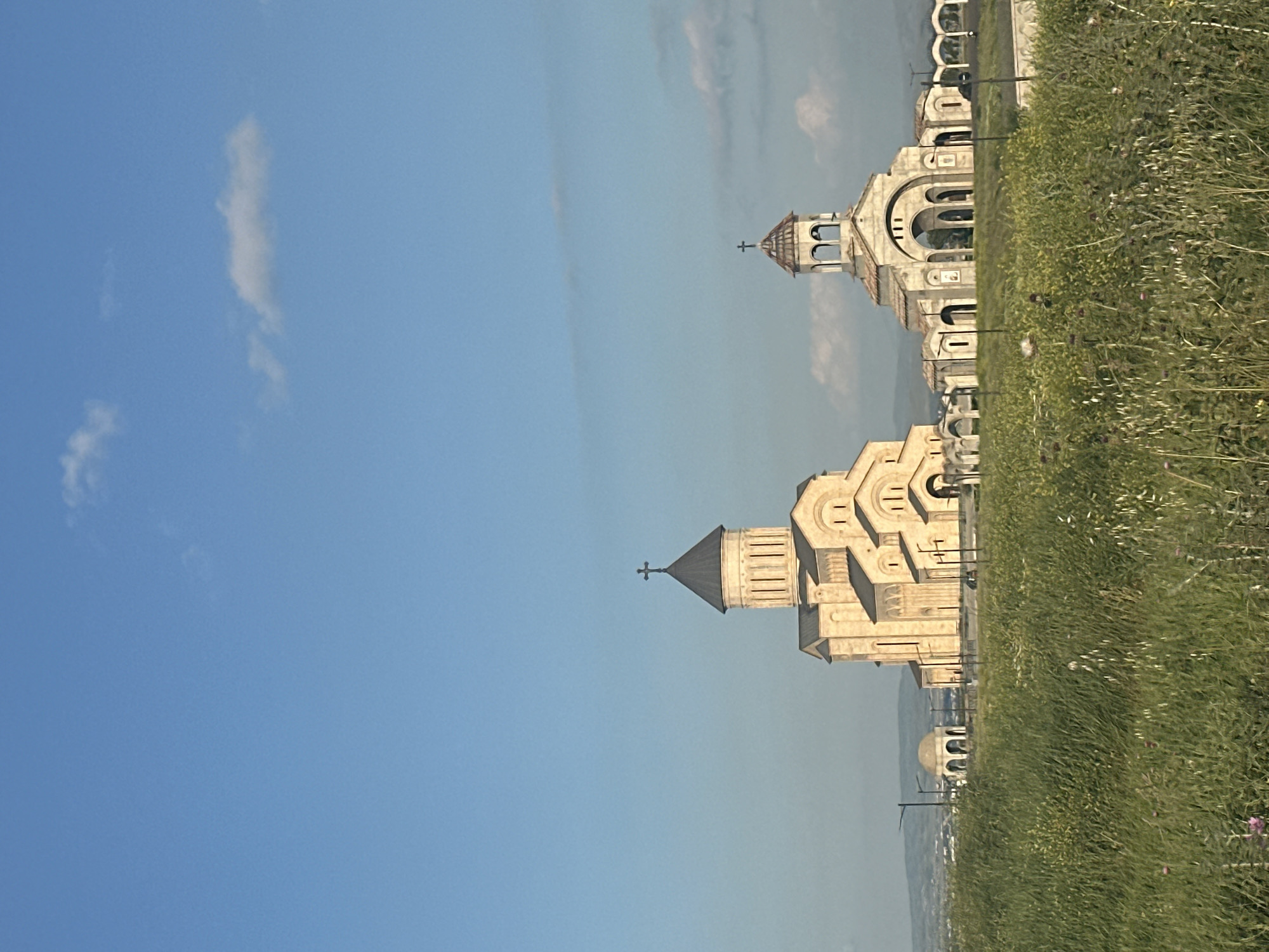 The image shows the Holy Trinity Cathedral of Tbilisi, Georgia, known as Sameba Cathedral. The cathedral is a prominent landmark situated on a hill overlooking the city. The photograph captures the cathedral from a distance, with a large grassy field in the foreground. The sky is clear blue with a few wispy clouds.
The main cathedral structure, with its golden domes and intricate architectural details, is the focal point of the image. To the left of the main cathedral, there is a smaller bell tower and a few other religious buildings. The surrounding area appears to be a public space, possibly a park or a religious complex, with a fence running along the perimeter.
The lighting suggests it is daytime, likely late afternoon or early morning, as the sun casts a warm glow on the cathedral's facade. The overall impression is one of grandeur and serenity, highlighting the architectural beauty of the cathedral and its picturesque setting.