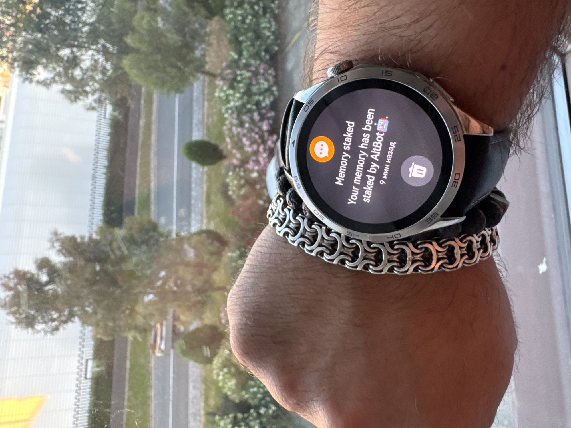 A person's wrist, adorned with a metal link bracelet and a smartwatch, is shown in the foreground. The smartwatch displays a notification stating "Memory staked Your memory has been staked by AltBot" with an icon of a robot and the text "9 мин назад" (9 minutes ago). The background is blurred, showing trees, foliage, a road, and a fence, suggesting an outdoor environment. The overall lighting indicates it is daytime. The scene is captured at a slight upward angle, focusing on the wrist and watch. No specific landmarks are identifiable that would pinpoint the exact location within Sirius, Russia.