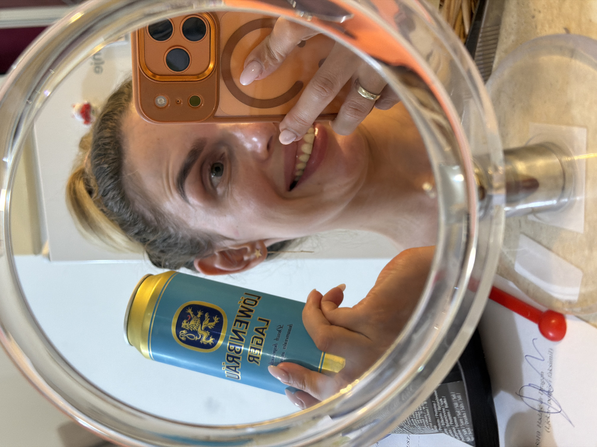 A woman is taking a mirror selfie in a room, reflecting herself and a can of Löwenbräu Lager. She is holding the beer can in her left hand and her orange iPhone in her right hand. She is smiling and looking at the camera. She has blonde hair pulled up in a bun, wearing a gold ring on her right ring finger and star-shaped earrings. In the background, there is a white wall and some objects on a desk, including what appears to be a paper with some writing. The setting does not provide enough information to determine the city of Budva, Montenegro.