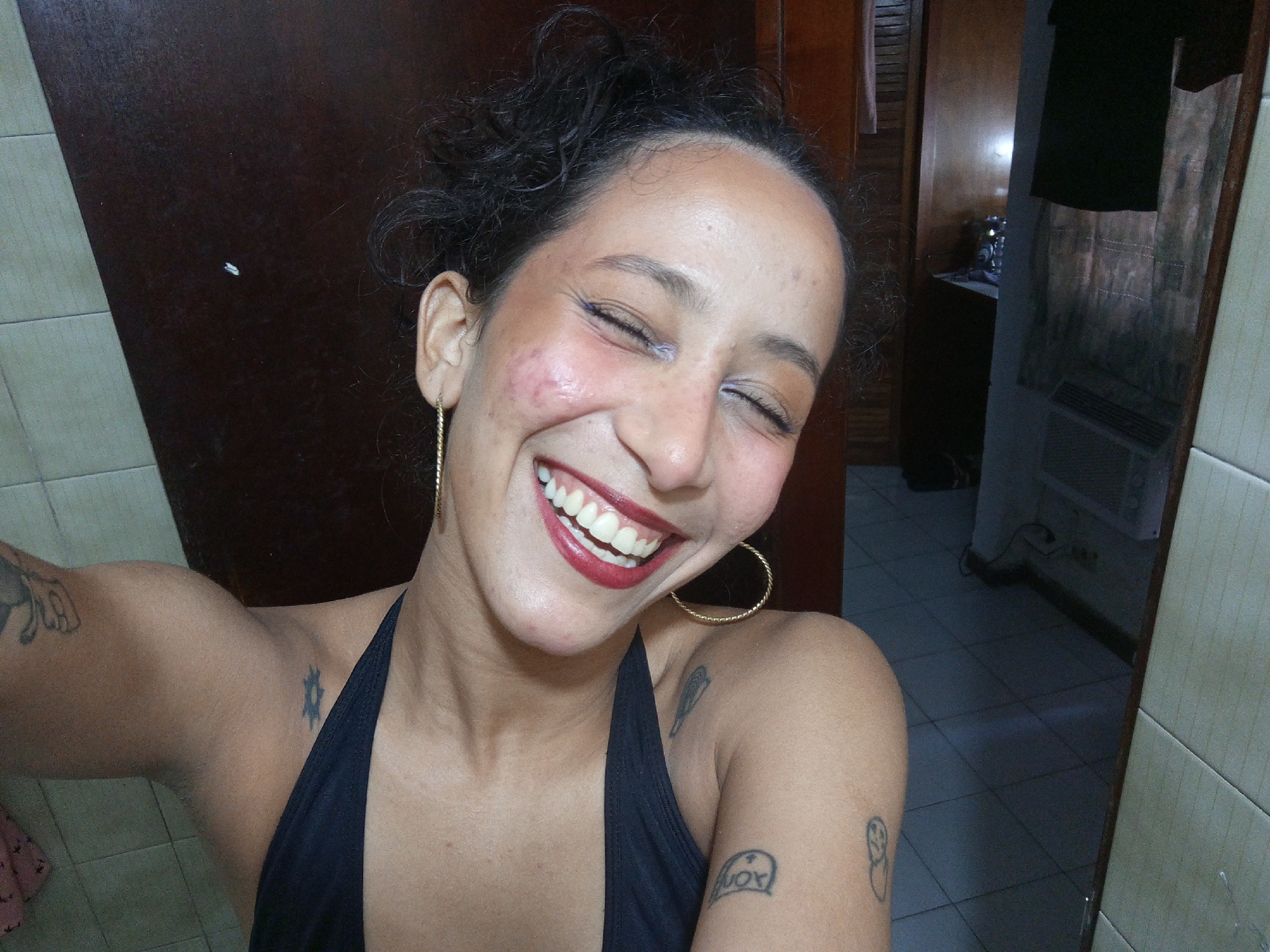 A smiling young adult woman with dark hair pulled back is taking a selfie indoors. She is wearing a black halter top and gold hoop earrings. Several small tattoos are visible on her arms and upper chest. The background includes a brown door and a glimpse into another room with light-colored tiled floor, a dark cabinet, and what appears to be an air conditioner unit. The location is likely in Porlamar, Venezuela. The image's details are insufficient to confirm the location with certainty.