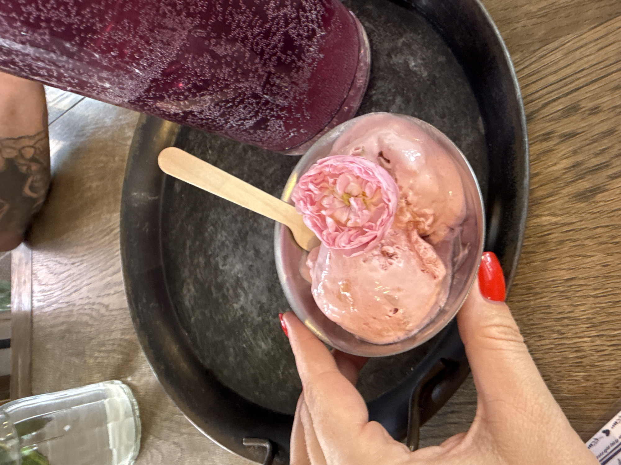 A close-up overhead shot shows a woman's hand with bright red nail polish reaching towards a small metal bowl filled with pink ice cream. The ice cream is topped with a delicate pink rose petal garnish. A wooden spoon rests in the bowl, partially submerged in the ice cream. The bowl sits on a dark metal tray, which in turn is placed on a textured wooden table. To the right of the tray, a tall glass bottle contains a dark purple, bubbly beverage. In the upper left corner, a partially visible clear glass bottle, possibly containing water, is also on the table. The background is blurred, suggesting an indoor setting. The scene is located in Natukhaevskaya, Russia.