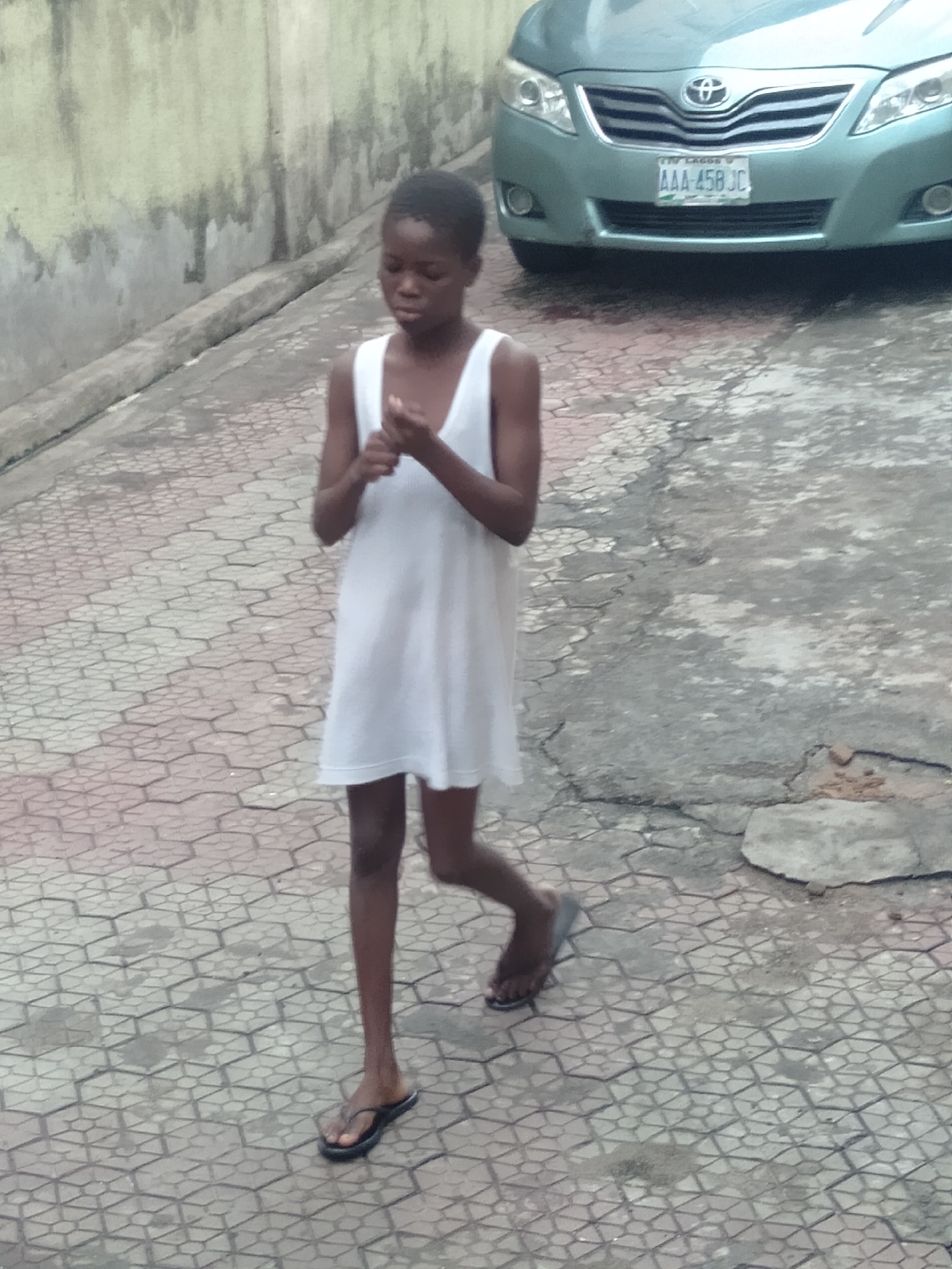 A young, dark-skinned person is walking on a paved surface next to a blue Toyota vehicle. The person is wearing a white dress and black sandals. The vehicle has a license plate number AAA-458 JC. A wall is visible in the background. The location appears to be an urban setting.