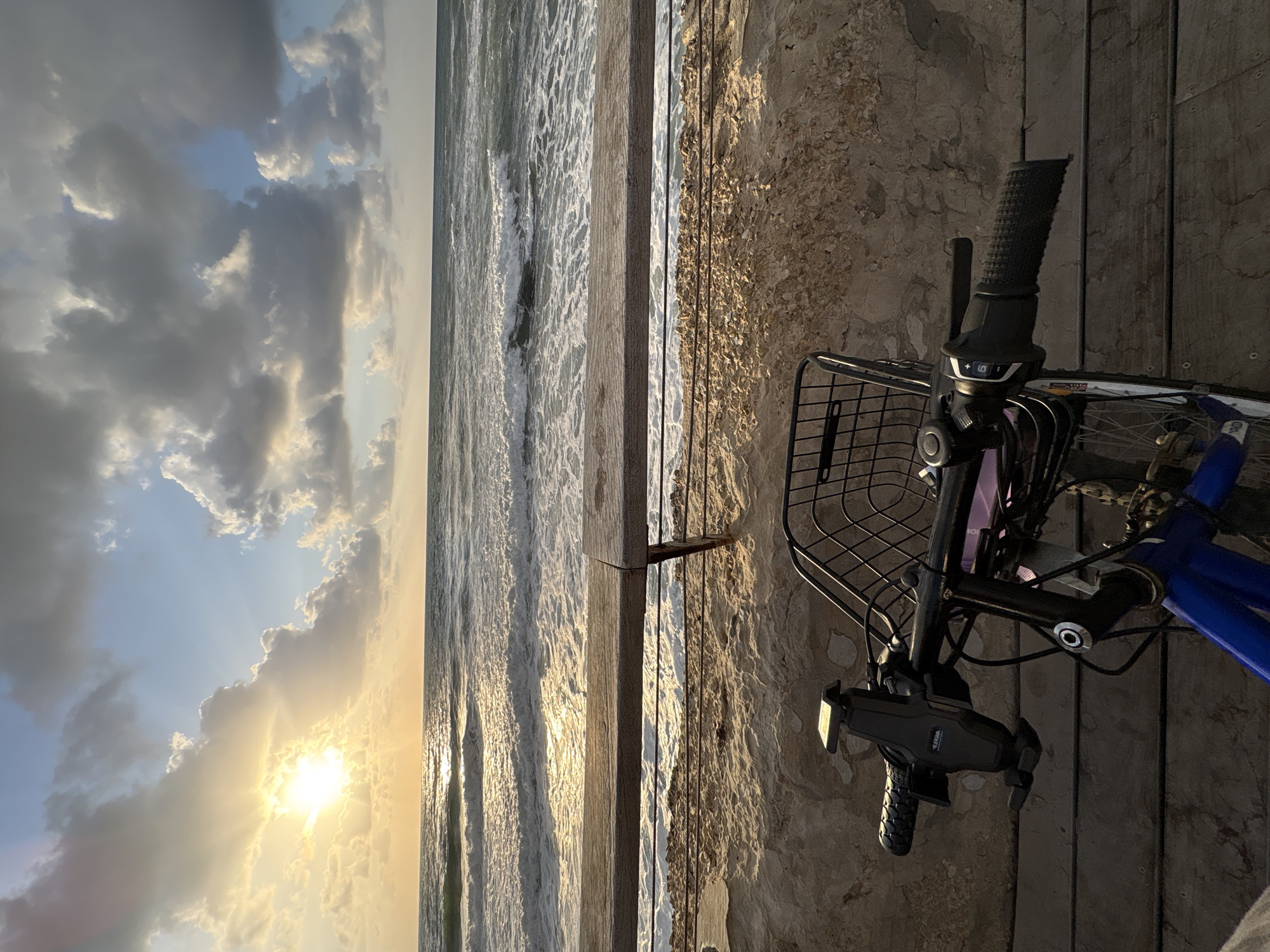 This image captures a serene coastal scene, likely at sunrise or sunset, viewed from a perspective that includes a bicycle in the foreground. Although the image is oriented vertically, it depicts a landscape where the horizon runs vertically through the center.
In the immediate foreground, positioned on the right side of the vertical frame (or lower right if viewed horizontally), is the front section of a bicycle. Key visible components include the black, textured grip of the handlebars, a gear shifter marked with '+ 6 -', a black wire mesh basket, and a black phone holder attached to the handlebars, which clearly displays the brand name "VOGU". Part of the blue bicycle frame and some wheel spokes are also visible. The bike rests on a textured concrete or stone surface, possibly a promenade, which features small embedded stones or shell fragments.
Separating the foreground from the sea is a narrow, weathered wooden beam or barrier. Beyond this, the midground is dominated by the sea, characterized by foamy waves breaking and reflecting the golden light from the sun. The water's surface shimmers with the sunlight.
The background, occupying the left side of the vertical frame (or upper left if viewed horizontally), showcases a dramatic sky filled with clouds. These clouds range in color from dark gray and blue to bright white and golden, illuminated by the low sun. The sun itself is visible through a break in the clouds, casting intense light rays and creating a strong glare. The sky transitions from a lighter blue in areas further from the sun to a warm, radiant golden hue near the horizon.
The overall atmosphere is peaceful and picturesque, capturing a moment of tranquility along a coastline, consistent with a setting like Tel-Aviv, Israel.
