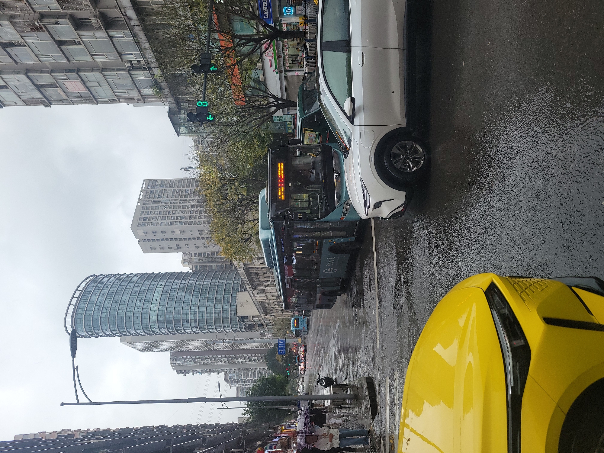 This image captures a rainy daytime street scene in Liangjiang, China, viewed from a slightly elevated perspective within a vehicle. The road is visibly wet, reflecting the overcast sky and the surrounding city lights.
In the foreground on the right, the bright yellow hood and fender of a car are prominent. Adjacent to it, a white SUV is stopped or parked. Further to the left, a green city bus, marked with the route number "113," occupies a lane, with Chinese characters visible on its side, likely indicating its route or transit company. Another blue bus and additional cars can be seen further down the street, amidst the flow of traffic.
On the left side of the street, pedestrians walk along the sidewalk, with some holding umbrellas, confirming the rainy weather. Various shopfronts line the street, including a "LAWSON" convenience store, and other businesses displaying Chinese characters. A striking, modern, curved glass building dominates the mid-ground on the left, standing out against more conventional multi-story buildings that extend into the background.
Traffic lights are visible at an intersection, displaying green directional arrows and a numerical countdown "8." Leafless or sparsely leaved trees line the sidewalk, and streetlights are also present. The scene depicts a bustling urban environment under wet, grey skies.