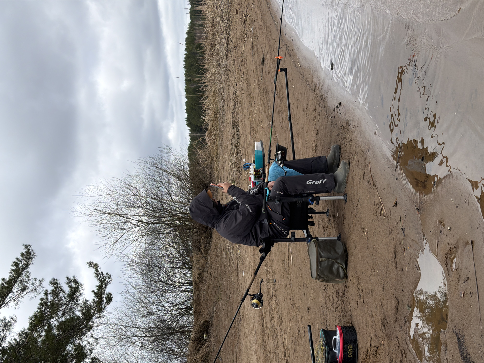 A person is sitting on a fishing chair on a sandy riverbank. They are wearing a black hooded jacket, black trousers with "Graff" printed on the leg, and green rubber boots. The person is holding a smartphone, presumably interacting with it. There are two fishing rods nearby. The immediate environment is a sandy riverbank with water flowing alongside. Further inland, there is dry vegetation and trees. The sky is overcast. The image does not contain sufficient information to confirm the location in the city of Chaykovsky, Russia.