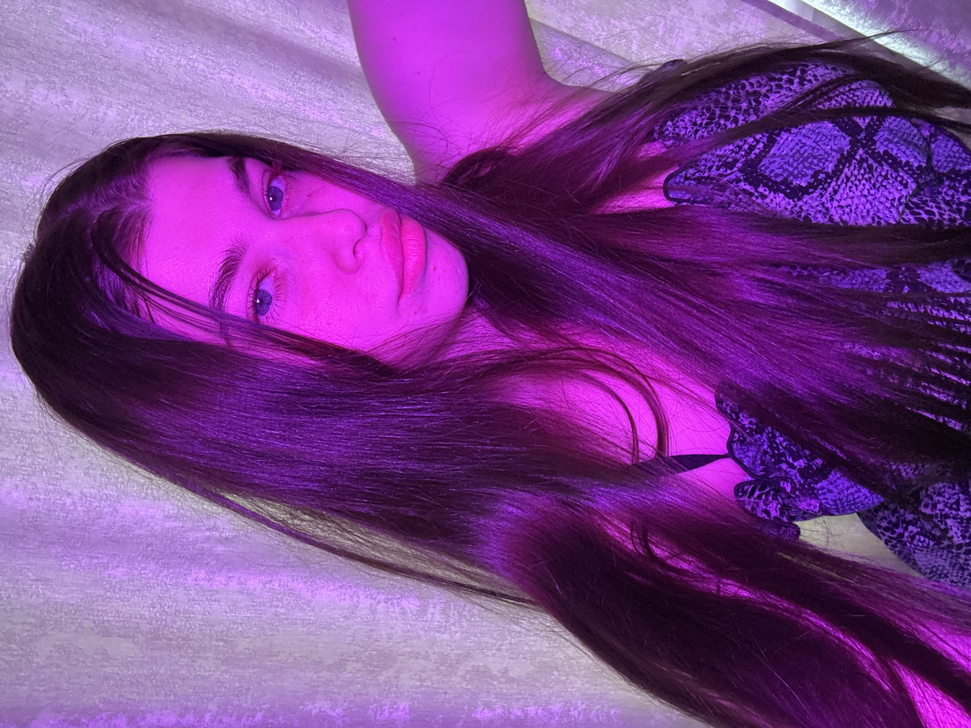 A woman with long brown hair lies on a textured white surface. Her face is illuminated with a purple light, emphasizing her blue eyes and pink lips. She wears a snake-print patterned top. The setting appears to be indoors, with an unspecified background. No information indicates the location is in Мещёры, Russia.
