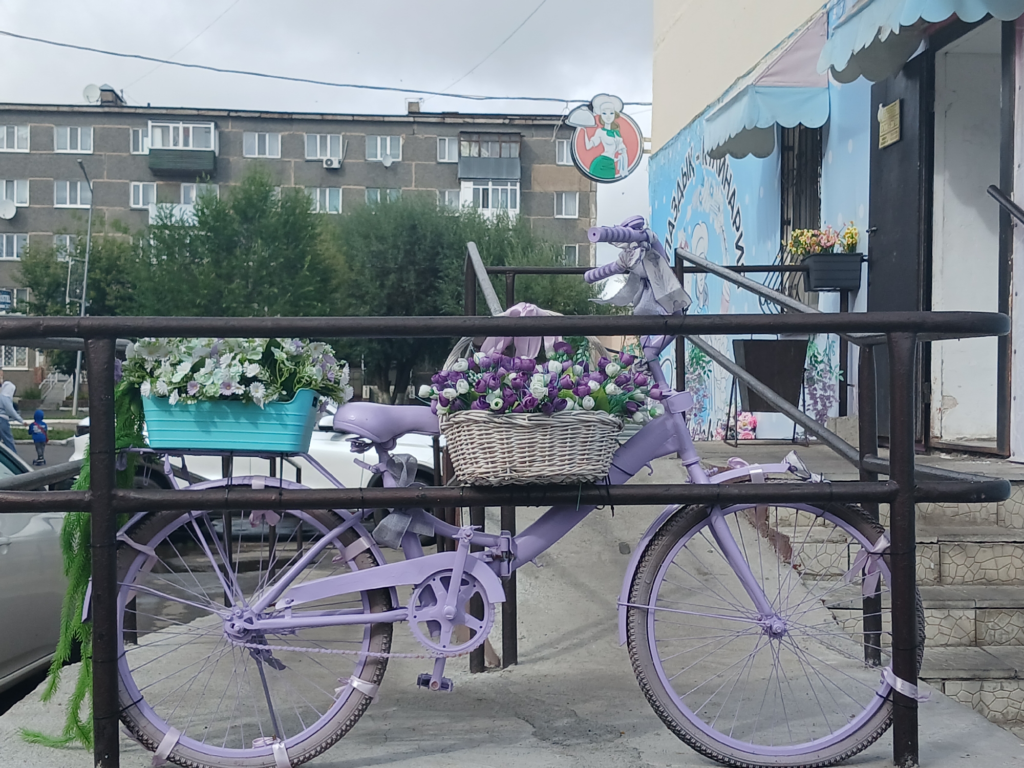 A lavender bicycle, adorned with floral arrangements in a turquoise basket and a woven basket on the handlebars, is parked in front of a building. The bicycle is secured by a dark metal railing. The building features light blue siding with a graphic of a woman in a chef's hat. A second, smaller planter with yellow flowers is attached to the building's facade. Behind the bicycle, a multi-story apartment building with gray concrete walls and numerous windows is visible. Trees with green foliage are scattered between the apartment building and the foreground. The sky is overcast. The scene appears to be an outdoor area in the city of Щучинск қаласы әкімдігі, Kazakhstan.