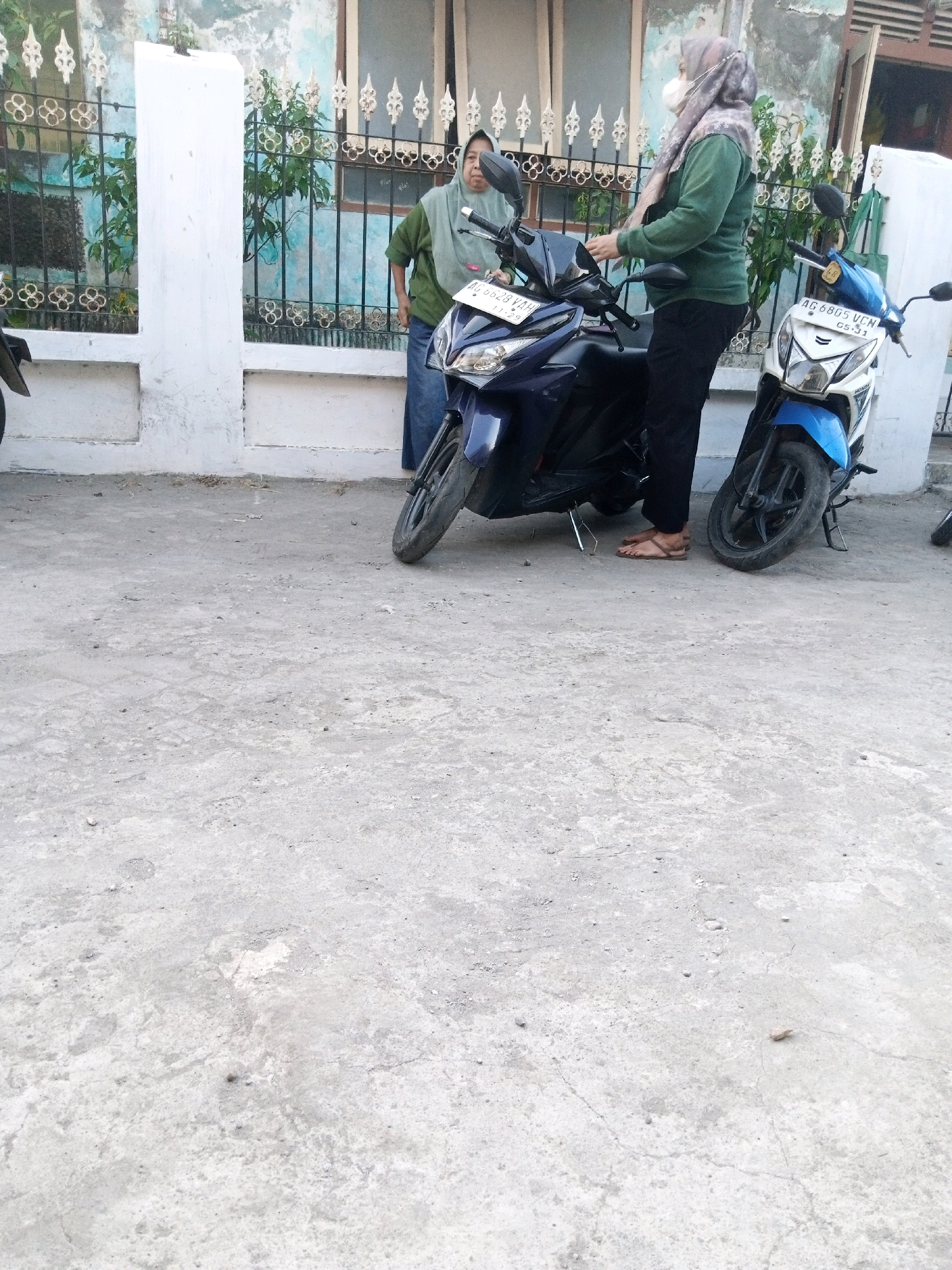 Two women are standing near motorcycles in front of a building in Sono Ageng, Indonesia. The woman on the right is wearing a green sweatshirt, black pants, and a gray hijab, and has a white mask on her face. She is standing next to a white and blue motorcycle. The woman on the left is wearing a green shirt and a blue skirt. She is standing next to a dark blue motorcycle. The building behind them is light blue with white trim and decorative black metal fencing. The ground in front of the building is concrete.