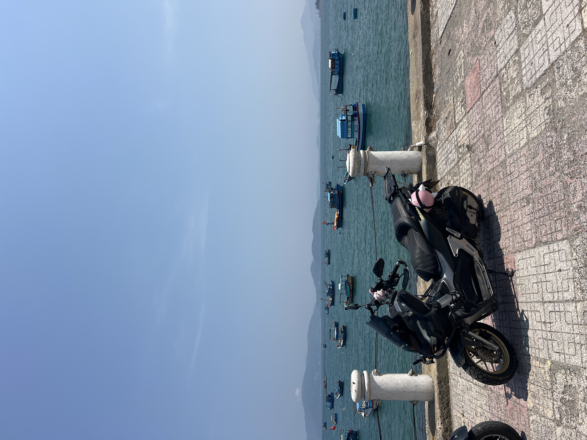 A medium shot shows a black motorcycle parked next to a stone barrier with several white cylindrical posts separating a tiled walkway from an ocean view in Bắc Nha Trang, Vietnam. The sky is a clear, light blue with minimal cloud cover. The ocean is a teal color, reflecting the sky. Numerous fishing boats, mostly blue and white, are scattered across the water. A mountain range appears faintly on the horizon. The walkway is made of light-colored, slightly weathered tiles with a grid pattern. The motorcycle is parked perpendicular to the barrier, facing towards the right side of the frame, with a pink helmet hanging on the handlebars. The scene is brightly lit, suggesting it is daytime.