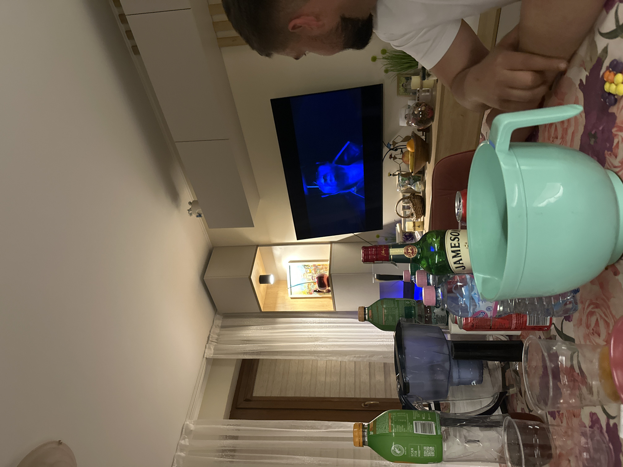 A man sits at a table inside what appears to be an apartment. The table is cluttered with various items including a large light blue bowl, bottles of water, and liquor bottles, including Jameson whiskey. Behind the man is a TV displaying a blue-toned image. Beyond the TV, a modern cabinet unit is visible, with an artwork displayed within the illuminated section. Curtains are visible on the right-hand side of the image, suggesting a window. The location appears to be an interior setting in an apartment but it's not possible to pinpoint the precise location in Plovdiv from just this one image.