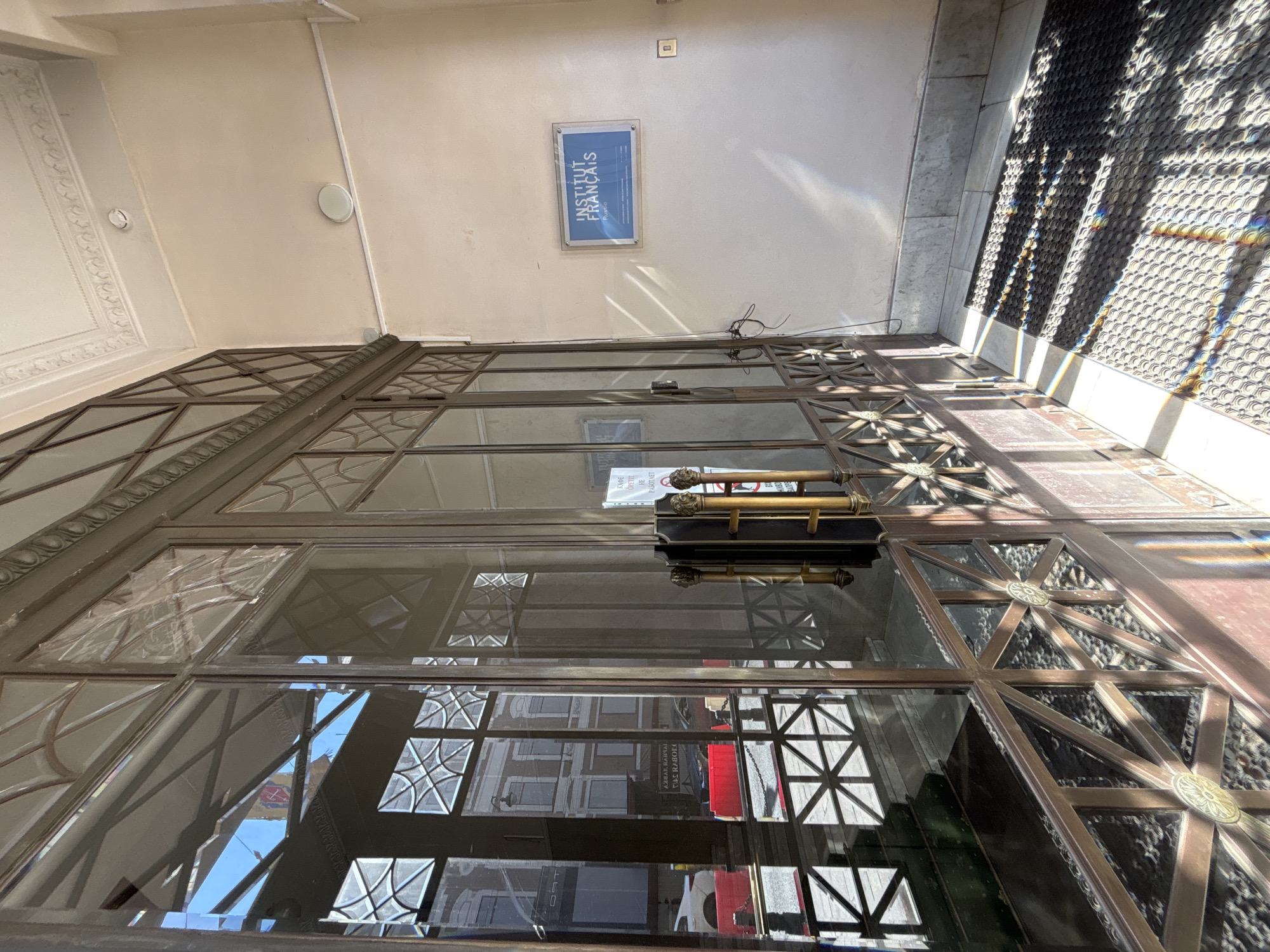 The image shows a partial view of the entrance to the Institut Français in Saint Petersburg, Russia. The entrance has a bronze-colored metal door with a grid-like pattern of windows. The reflection in the glass shows buildings across the street, suggesting a location in an urban area. A sign for the Institut Français is visible on the wall above the door. The wall is painted a light beige color. To the right of the entrance is a textured wall with a sunlight pattern.