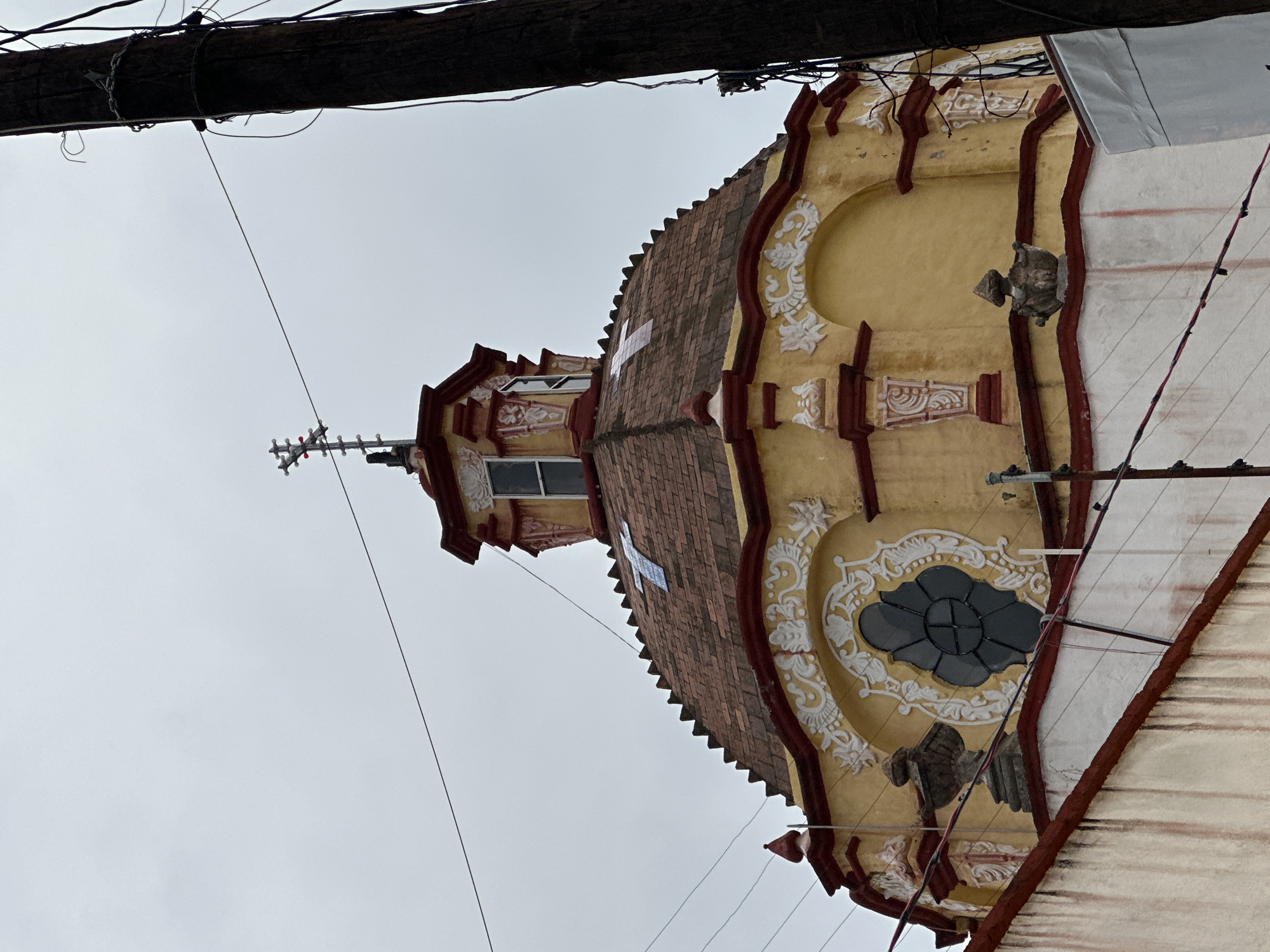 An upward, rotated view shows the ornate dome of a church, situated in San Miguel Zinacantepec, Mexico. The dome features yellow stucco walls, red trim and a brown tile roof with white decorative flourishes. Two sculpted gargoyle-like figures sit on either side of the dome. A cross tops the structure. Power lines and a utility pole cut across the top of the image. The sky is overcast.