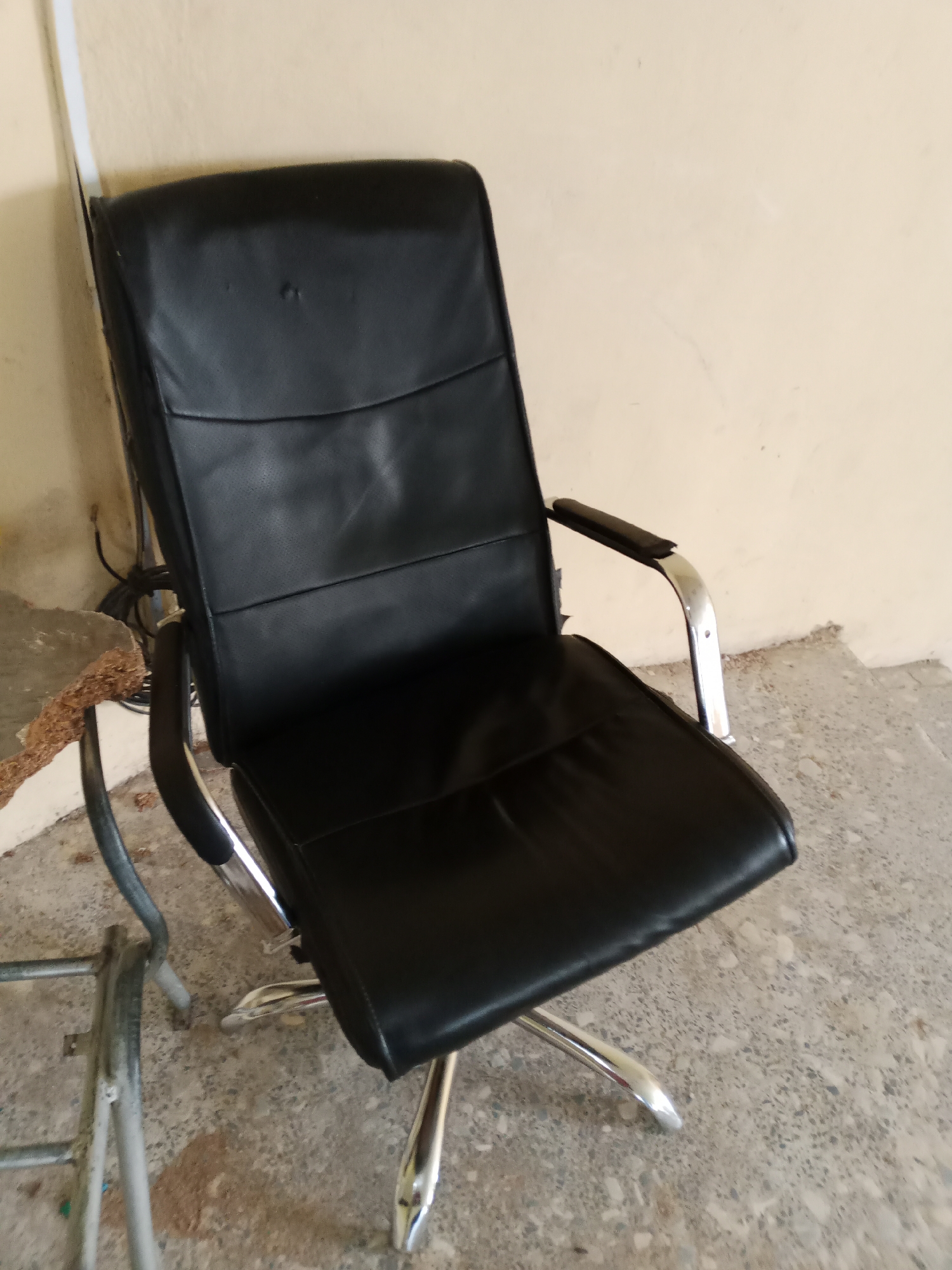 A black leather office chair with chrome accents sits on a speckled gray concrete floor in what appears to be an unfinished or outdoor area. The chair has a high back with horizontal stitched panels and armrests. To the left of the chair, a portion of a damaged, light-colored wall is visible, with some electrical wiring coiled on the floor. A rough, brown object, possibly a piece of damaged wood or insulation, lies on the floor in front of the wall. The background wall is painted a pale yellow. The scene suggests an urban or semi-urban setting, potentially in Jalingo, Nigeria, given the context. There are no people present in the image.