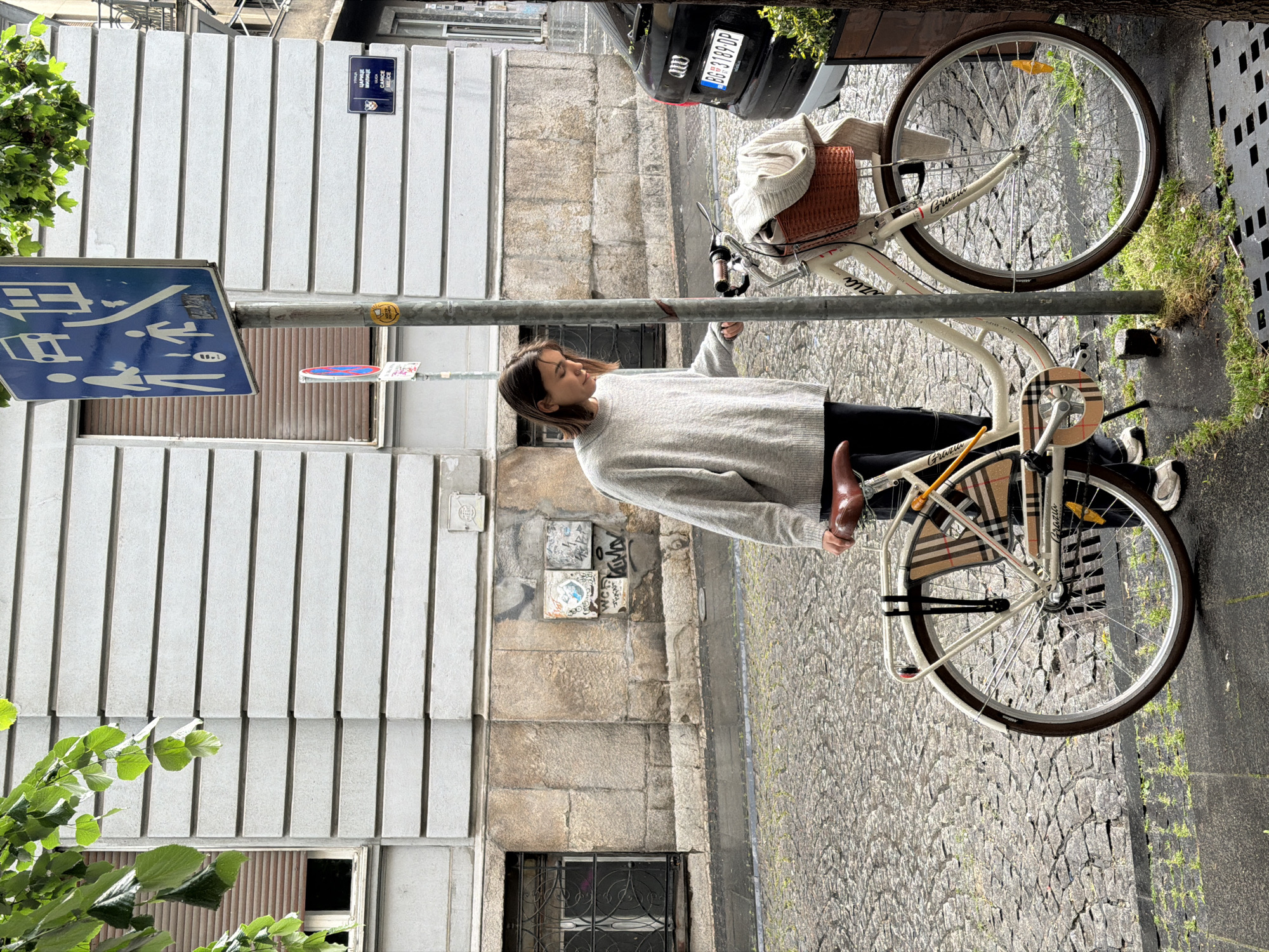 A young woman is standing next to a white bicycle secured to a metal pole on a cobblestone street in Belgrade, Serbia. The woman wears a light gray sweater and black pants, holding onto the pole with one hand. The bicycle has a wicker basket on the handlebars and a plaid pattern on the rear fender. The street is bordered by a building with a white, vertically-paneled facade and a lower section of stonework. A street sign is attached to the metal pole, as well as a street name sign reading "Царице Милице" ("Carice Milice"). A black car with a license plate is parked nearby. Green foliage is visible on either side of the image.