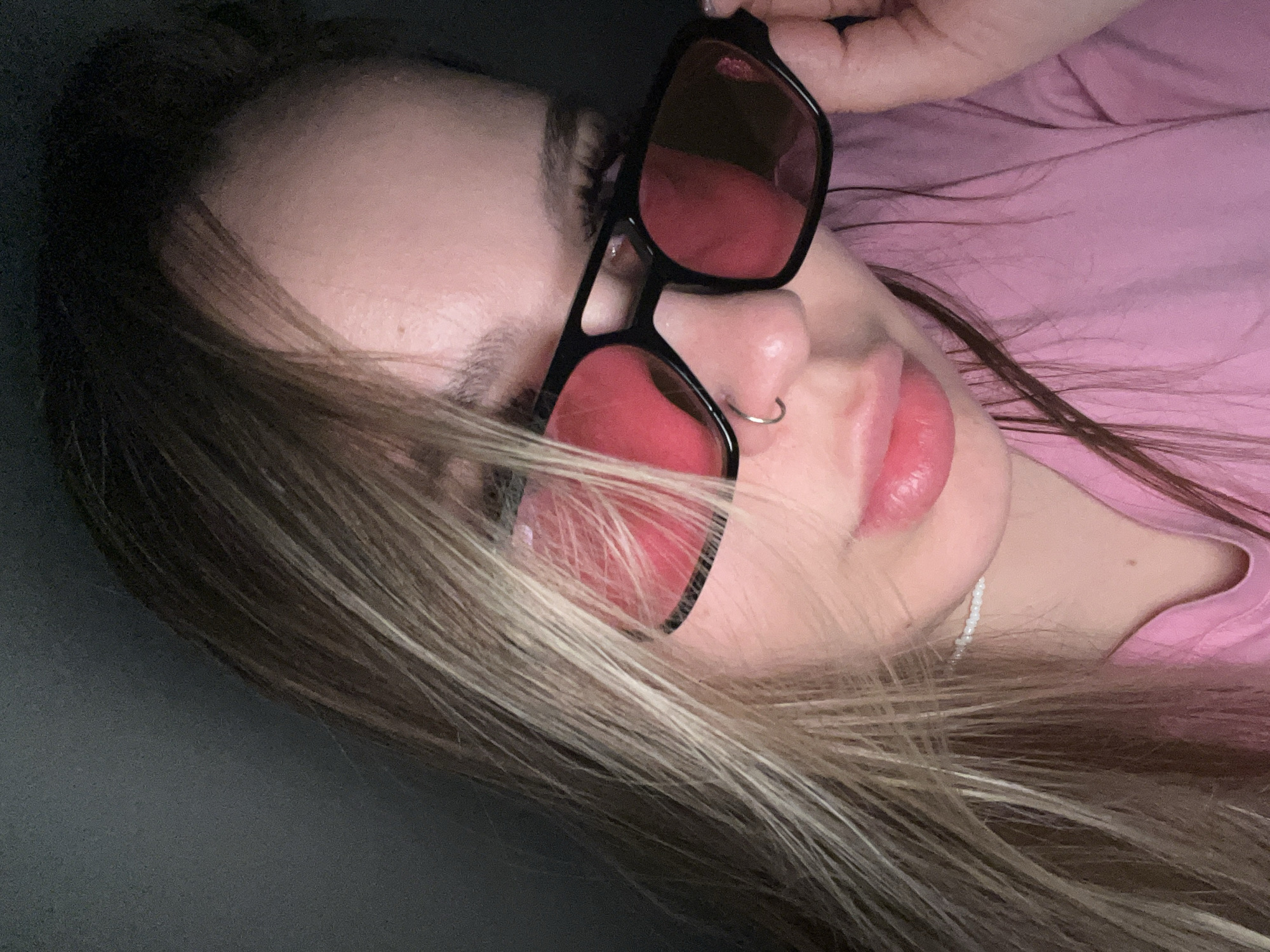A woman with blonde hair and wearing a pink shirt is seen from a low angle. She has a nose ring and is wearing black sunglasses with red lenses. The background is blurry and dark. The image was not taken in Krasnodar, Russia.