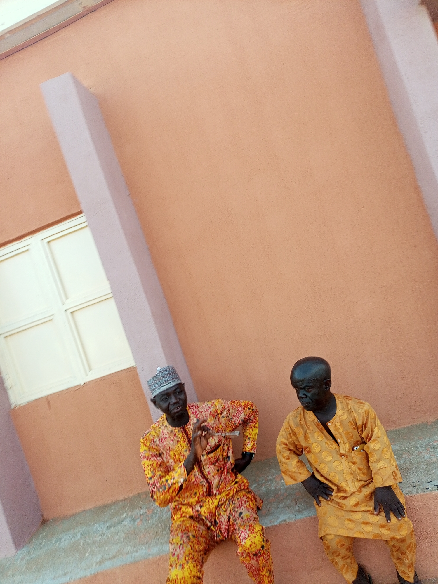Two men sit side-by-side on a concrete step in Tempure, Nigeria. The man on the left wears a patterned yellow and red outfit with a white and gray cap. The man on the right wears a gold-colored outfit. Both men are looking towards the left. Behind them is a peach-colored wall with a white window frame and a light pink pillar.