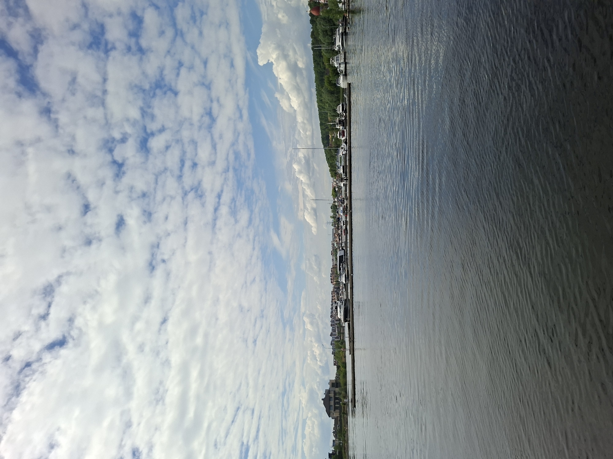 This image captures a view of the Kazanka River in Kazan, Russia. The scene is dominated by a wide expanse of dark, rippling water in the foreground, reflecting the sky. In the middle ground, a long pier juts out into the river, lined with numerous moored yachts and boats of various sizes. The pier area is busy with docked vessels, suggesting a marina or yacht club. Beyond the pier, the background reveals a shoreline with lush green trees and buildings. On the left side of the background, a large, modern house is visible. Further along the shoreline, a cluster of contemporary buildings and structures indicates urban development. The sky above is a mix of blue sky and scattered white clouds, creating a dynamic atmosphere. The overall impression is one of a pleasant day at a waterfront location, likely a leisure area within or near Kazan.
